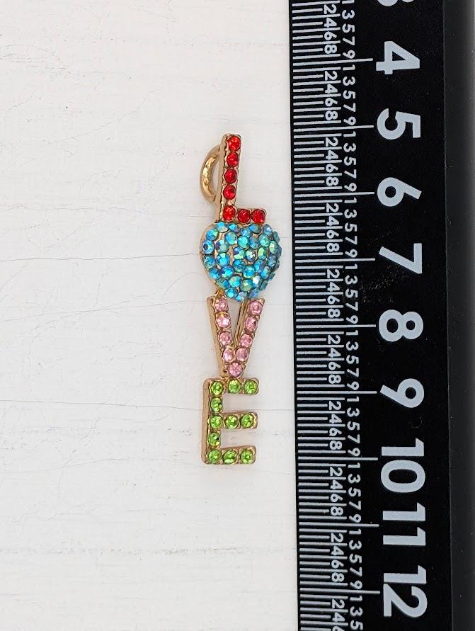 A colorful pendant or brooch with the word "LOVE" written on it, made of various materials including gemstones and metal.