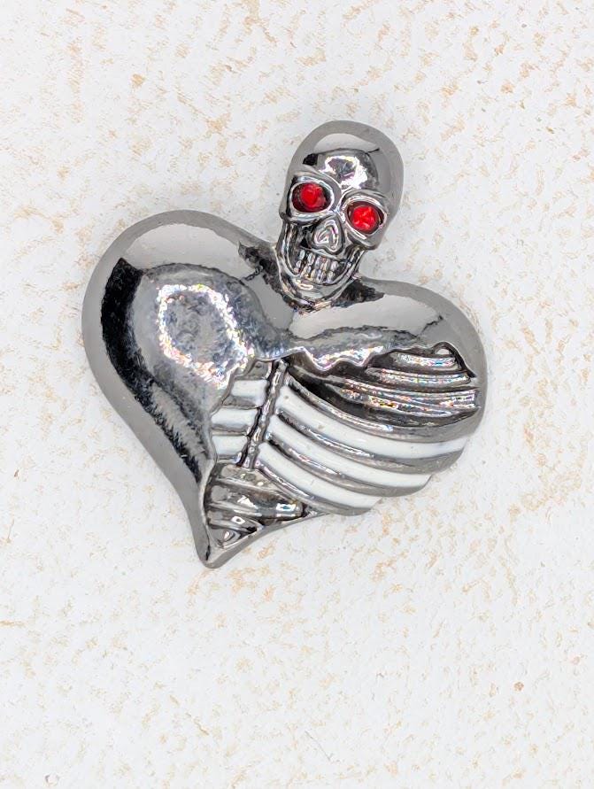 A silver metal heart-shaped brooch with a skull design on it, featuring red eyes.