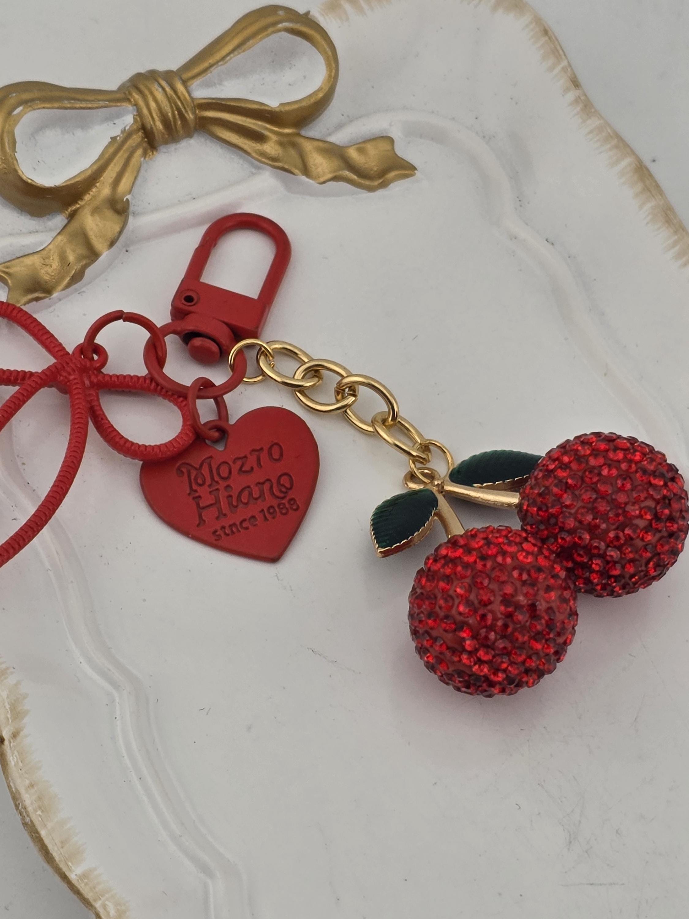 a keychain with a red heart-shaped charm and two red cherries, along with a red ribbon and a gold bow.