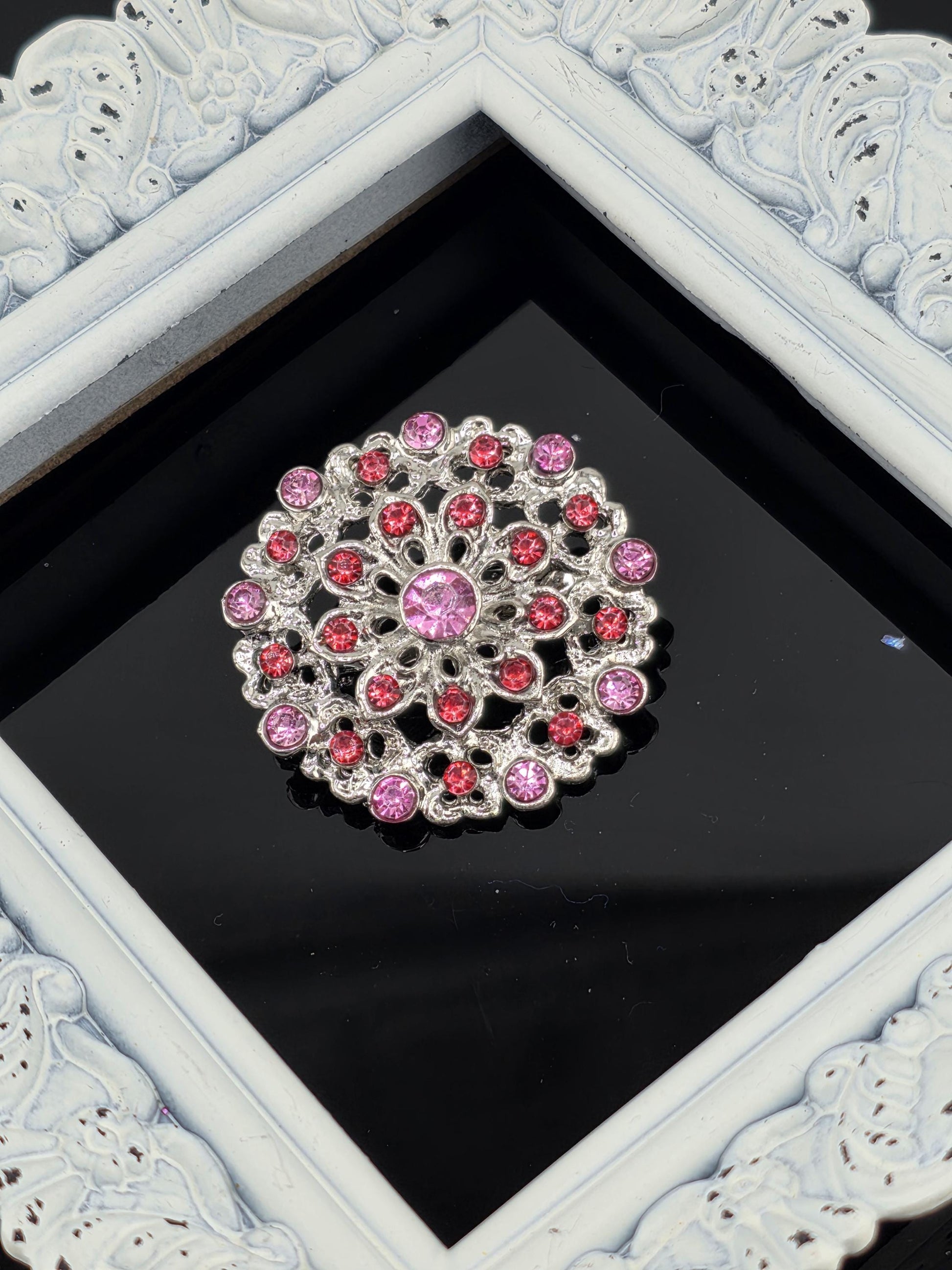 A decorative brooch with a floral design and pink and red gemstones is displayed against a black background, framed by a white ornate frame.