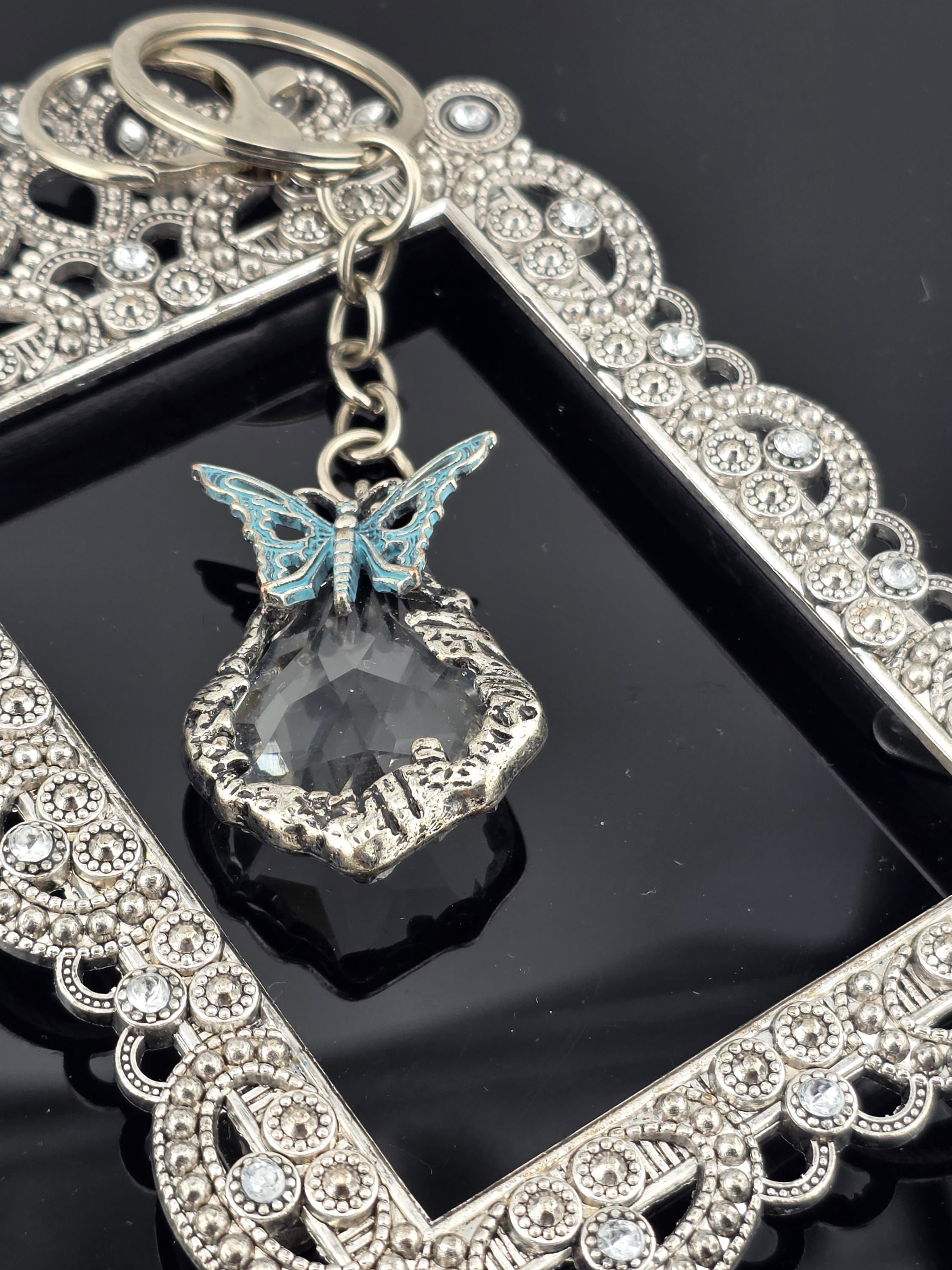 A silver-colored keychain with a clear gemstone and a butterfly-shaped charm is displayed against a black background.
