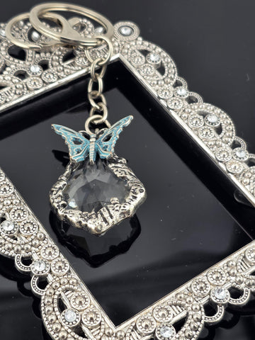 A silver-colored keychain with a clear gemstone and a butterfly-shaped charm is displayed against a black background.