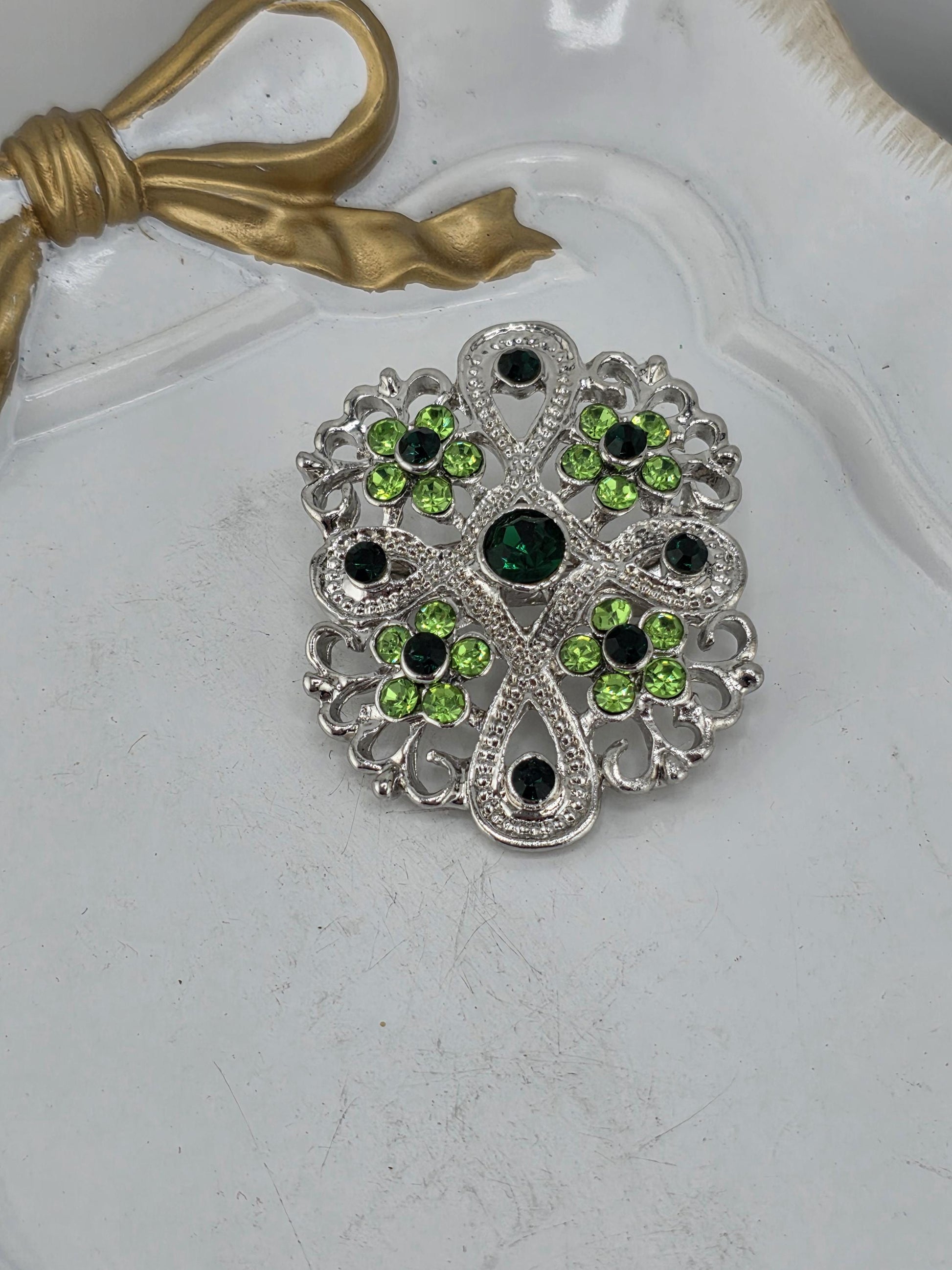 A decorative brooch with a floral design and green gemstones is displayed on a white surface.