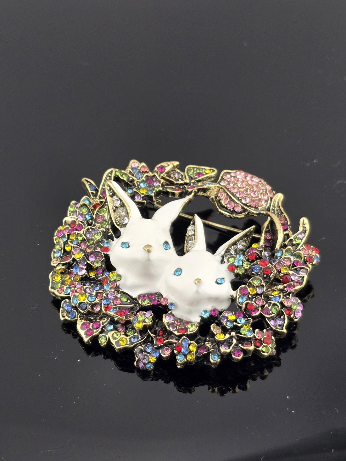 A decorative brooch with two white bunny heads, surrounded by a colorful floral wreath and a gold bow.