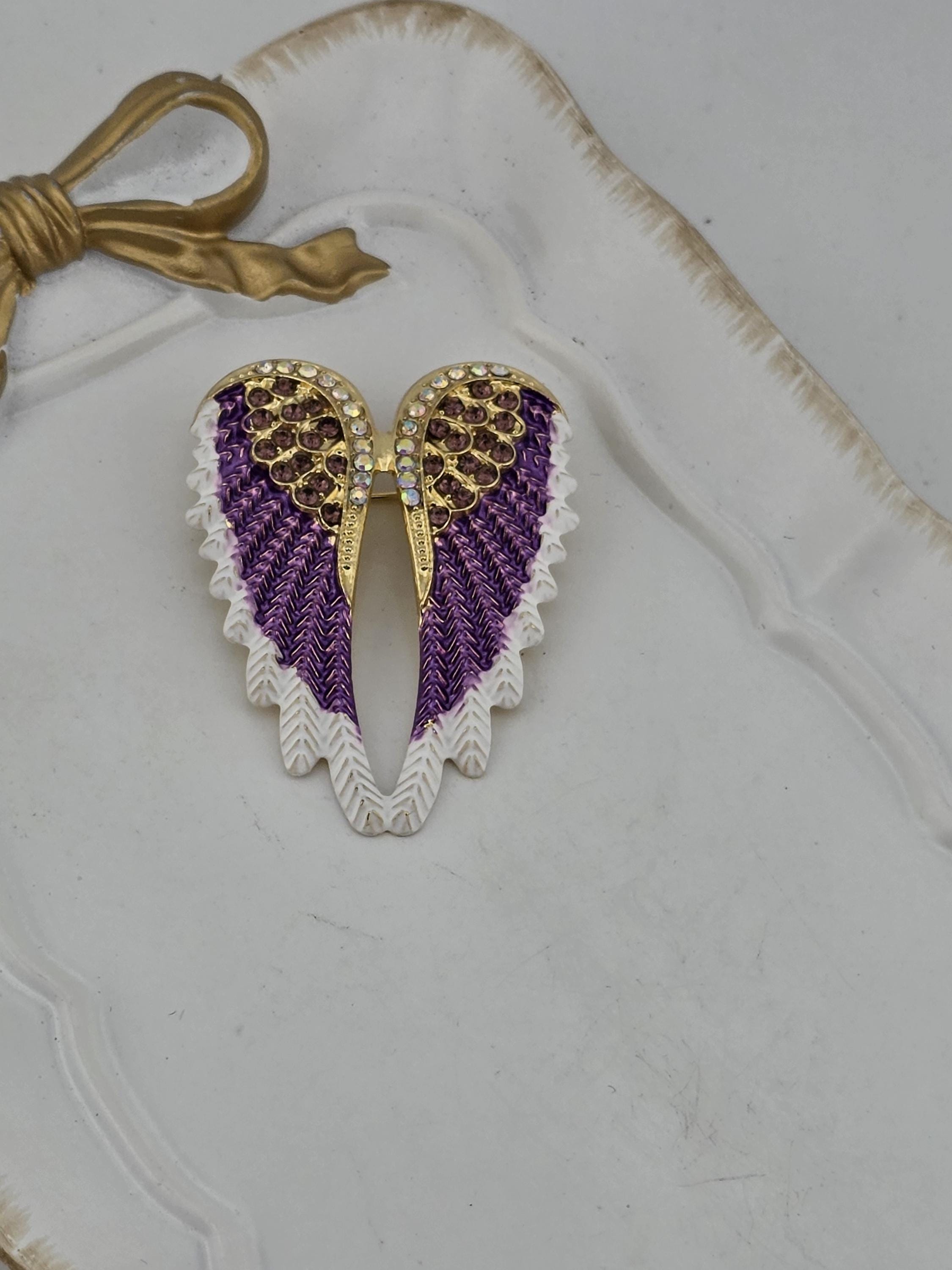 A decorative wing-shaped brooch with purple and gold accents, placed on a white surface.