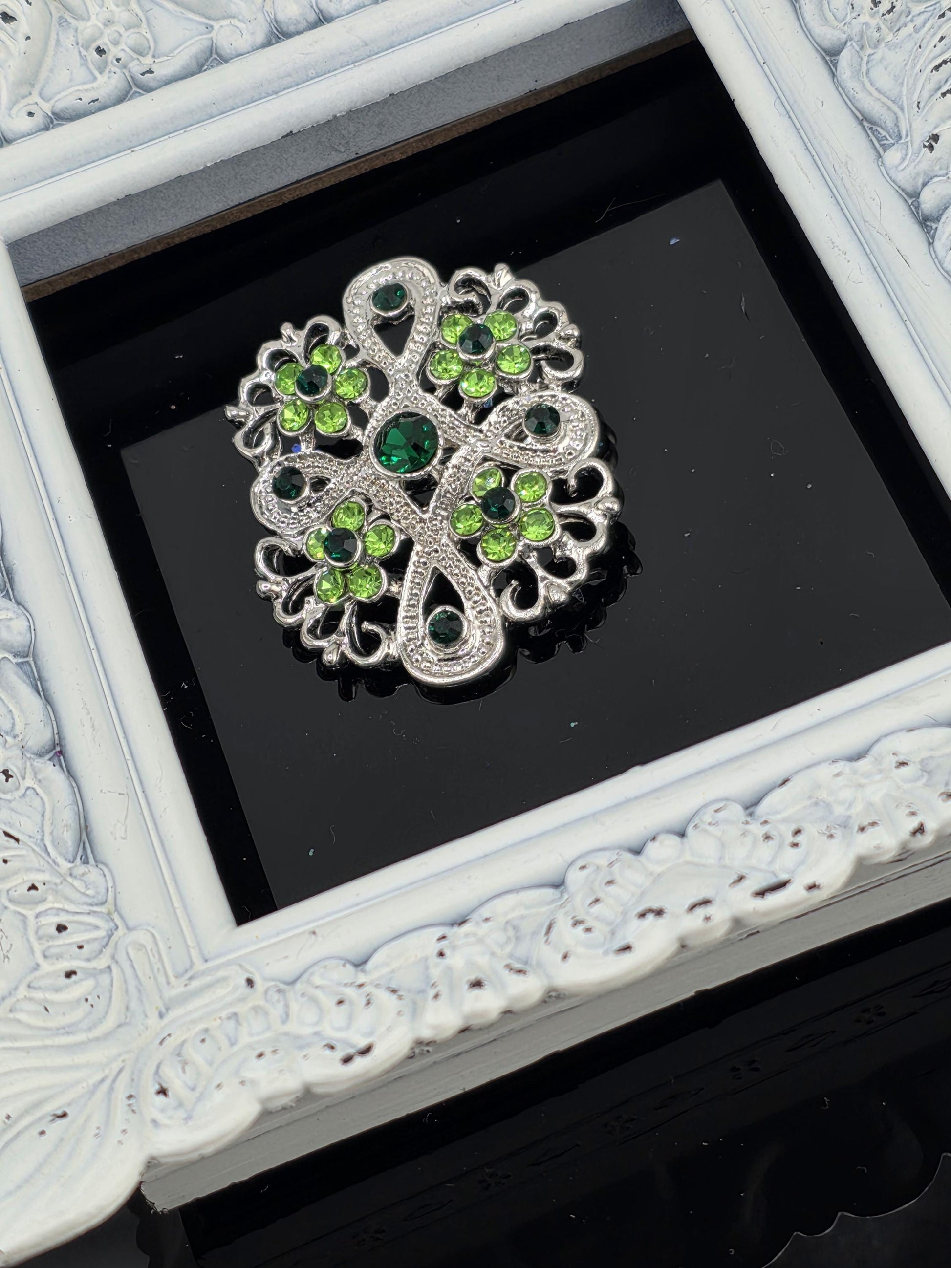 A decorative brooch with green and silver gemstones is displayed in a white ornate frame against a black background.