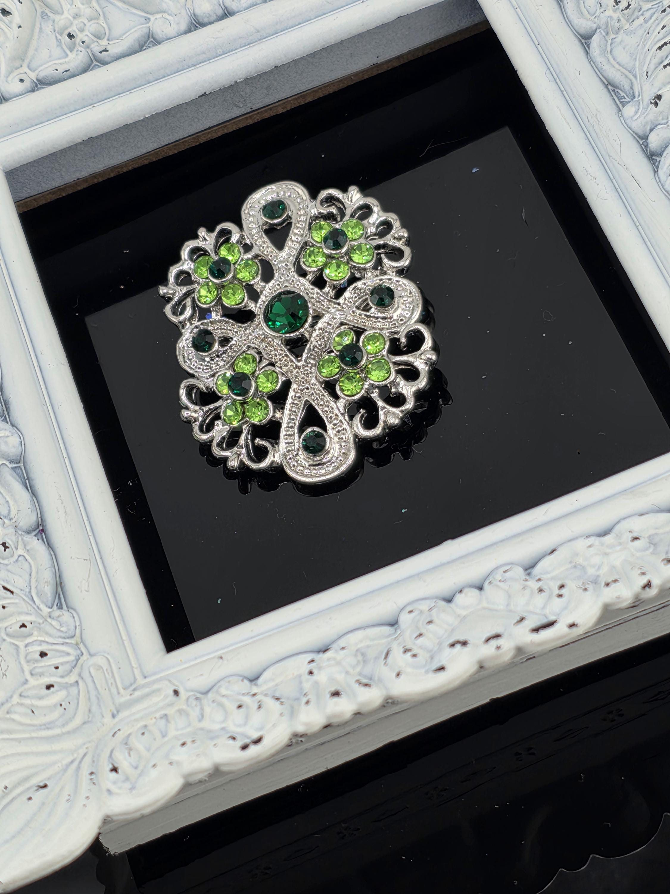 A decorative brooch with green and silver gemstones is displayed in a white ornate frame against a black background.