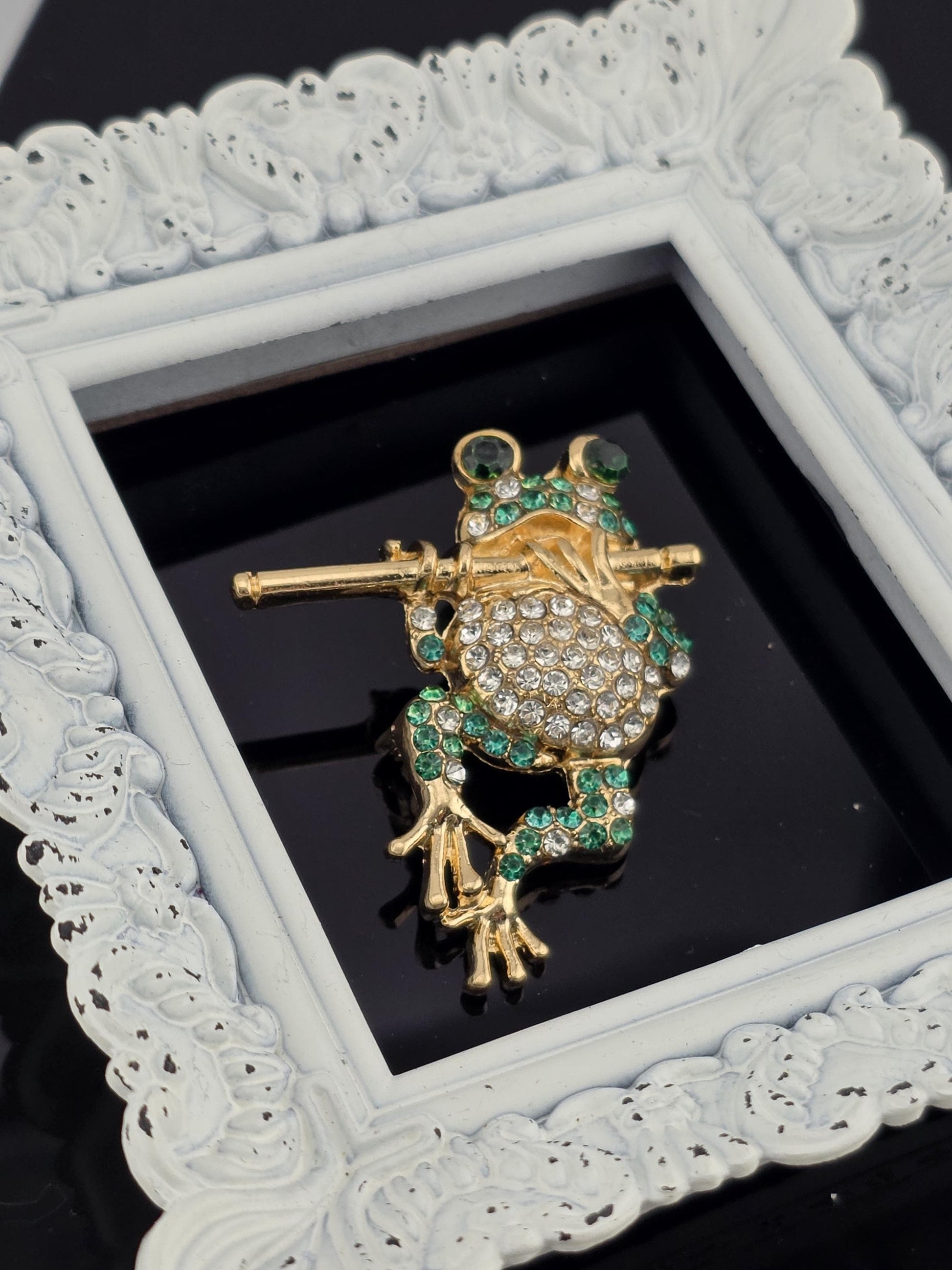 A decorative gold and green frog brooch with sparkling gemstones is displayed in a white ornate frame.