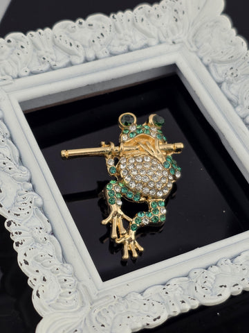 A decorative gold and green frog brooch with sparkling gemstones is displayed in a white ornate frame.