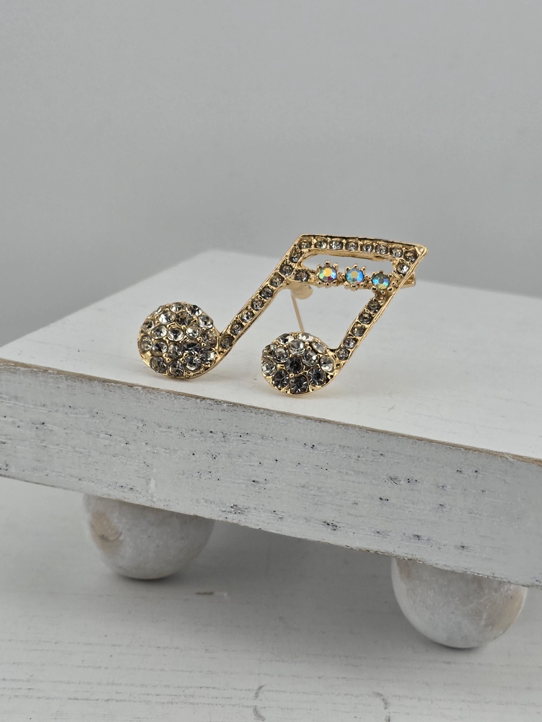 A gold-colored pin adorned with rhinestones in the shape of a musical note, resting on a white surface.