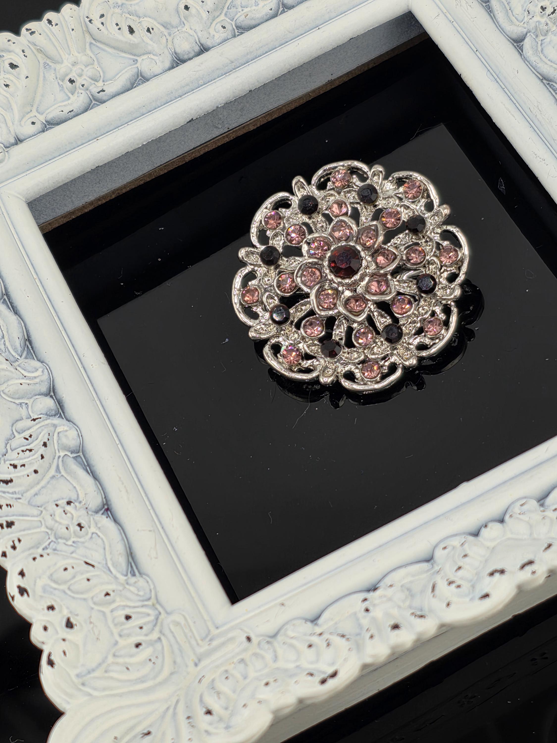A decorative brooch with a floral design and a central gemstone is displayed in a white ornate frame against a black background.