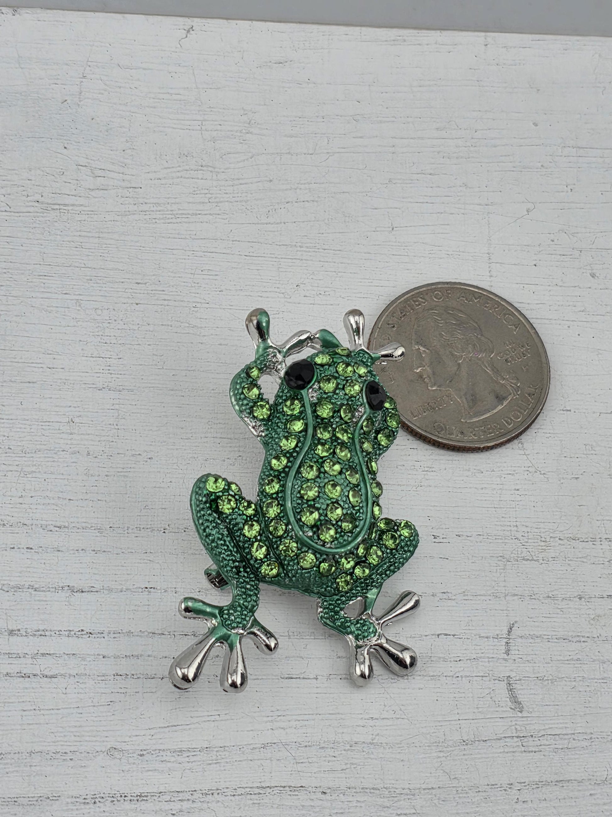 A green frog brooch with sparkling rhinestones is placed next to a silver coin on a white surface.