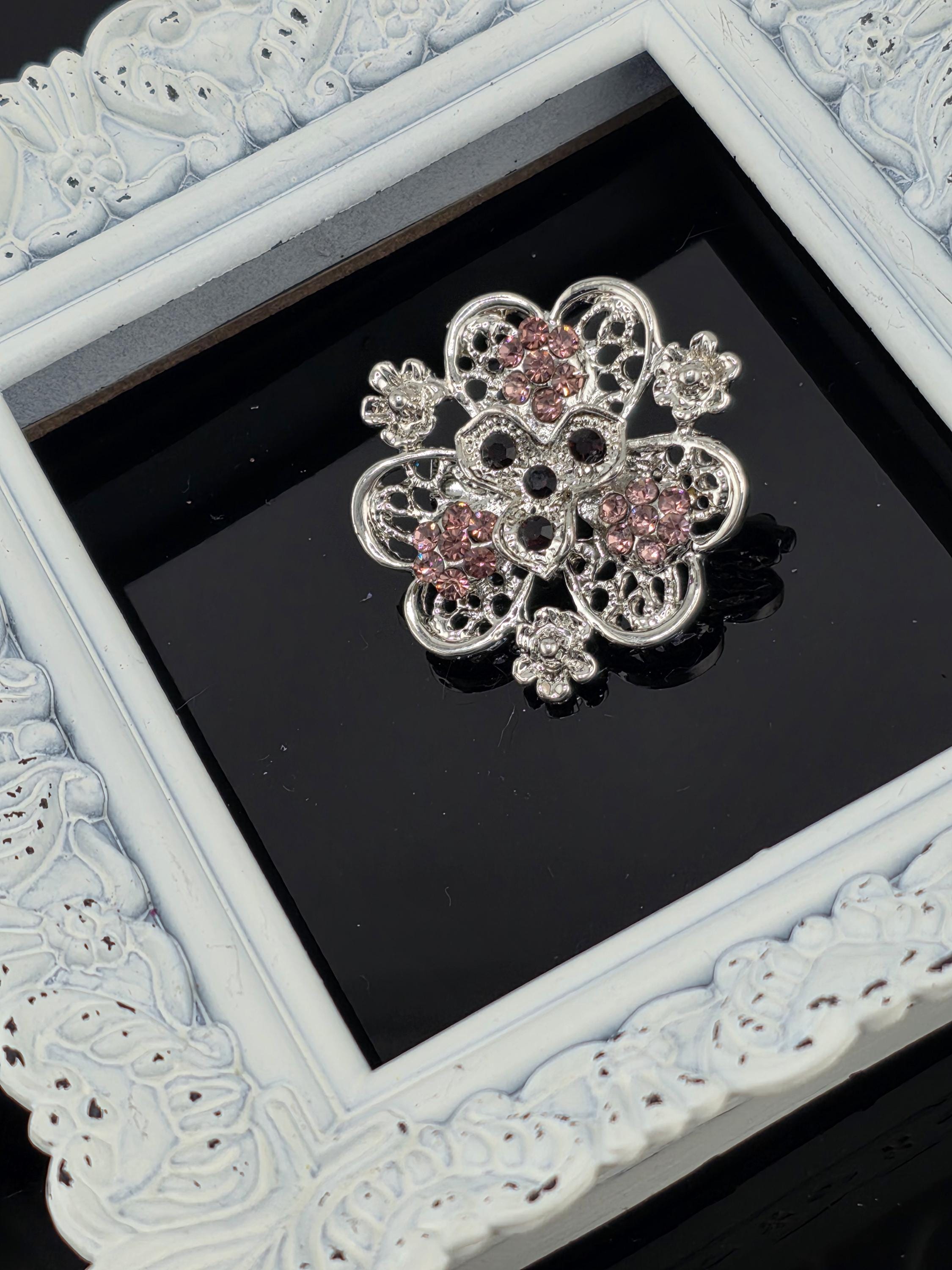 A decorative silver brooch with pink gemstones is displayed in a white ornate frame against a black background.