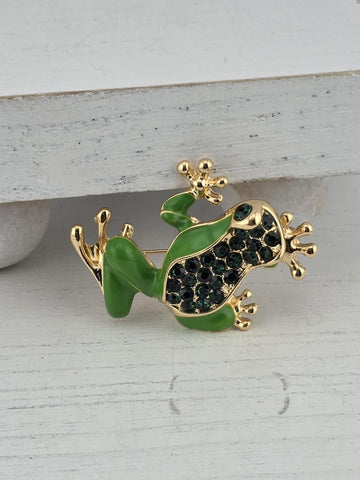 A green frog-shaped brooch with gold accents and small black gemstones is displayed against a white background.