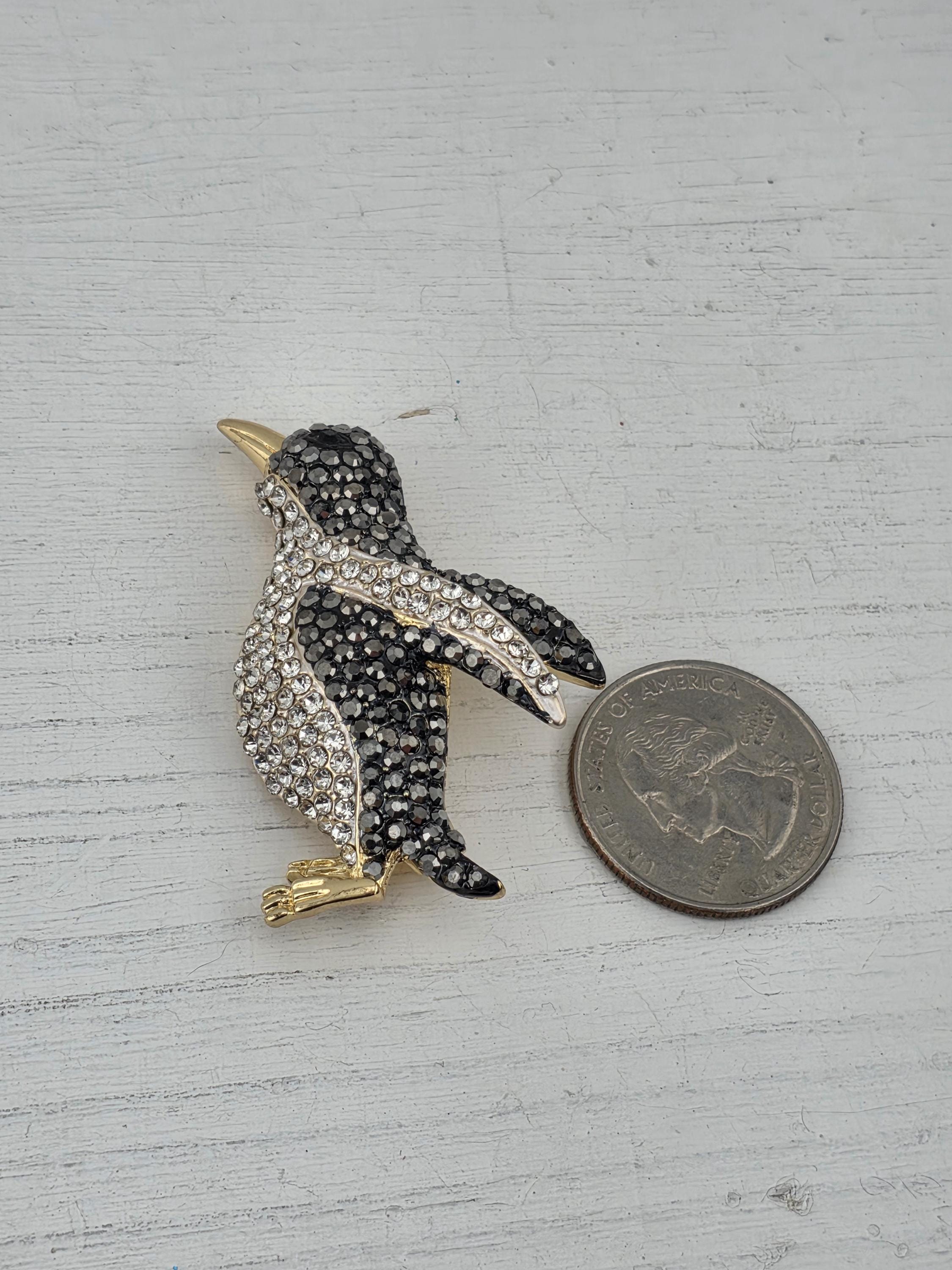 A shiny, black and white penguin brooch with sparkling rhinestones is placed next to a United States quarter on a white surface.