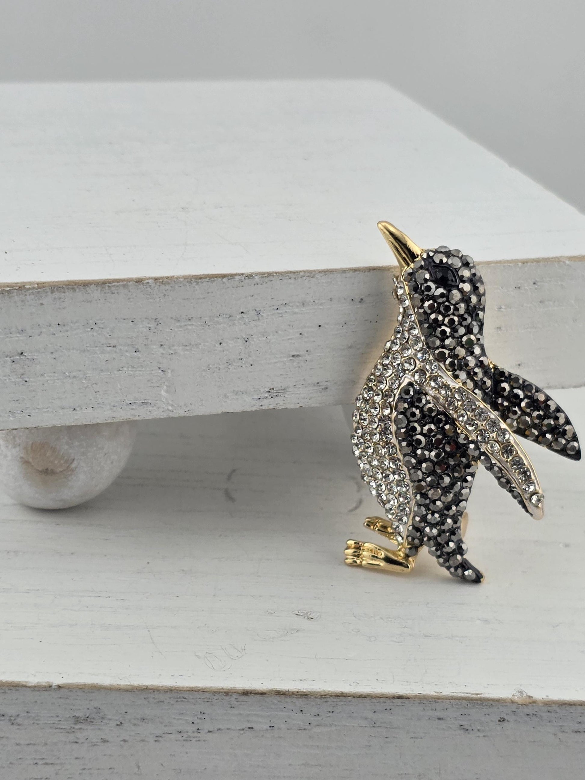 A decorative bird brooch with a sparkling black and white design, featuring a gold beak and feet, is displayed on a white surface.