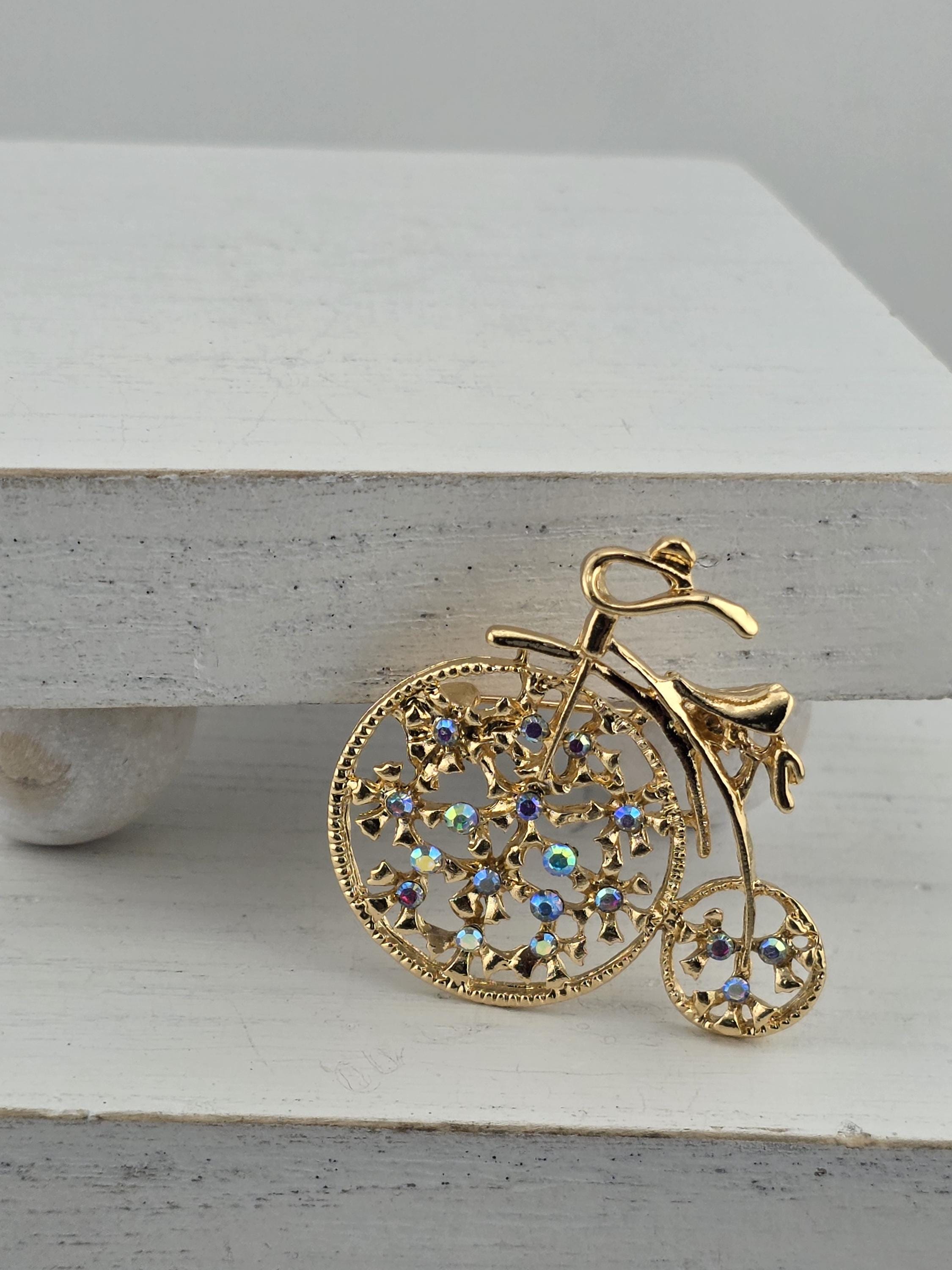 A decorative gold brooch in the shape of a bicycle, adorned with colorful gemstones, is displayed against a white background.