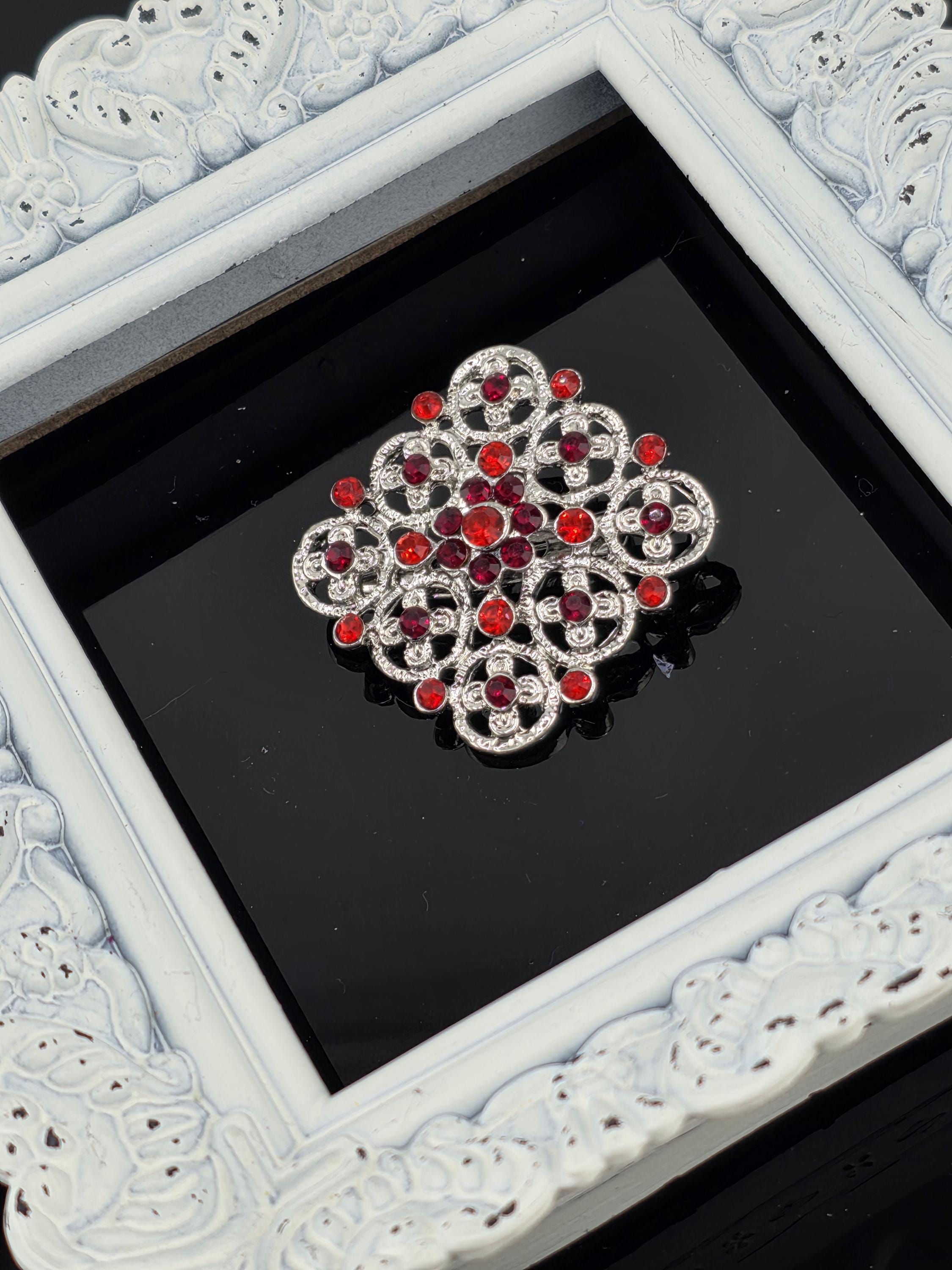 A decorative brooch with a floral design and red gemstones is displayed in a white ornate frame against a black background.