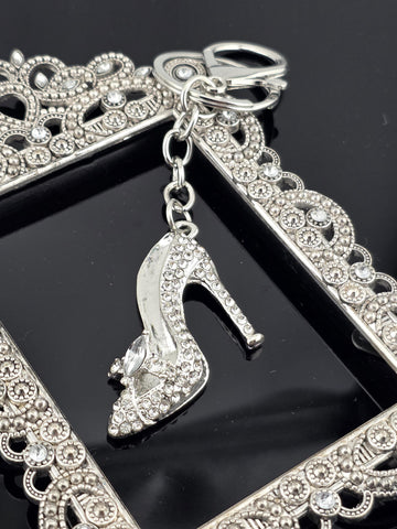 A close-up view of a silver-colored high heel shoe with rhinestones, attached to a decorative frame with a scalloped edge.
