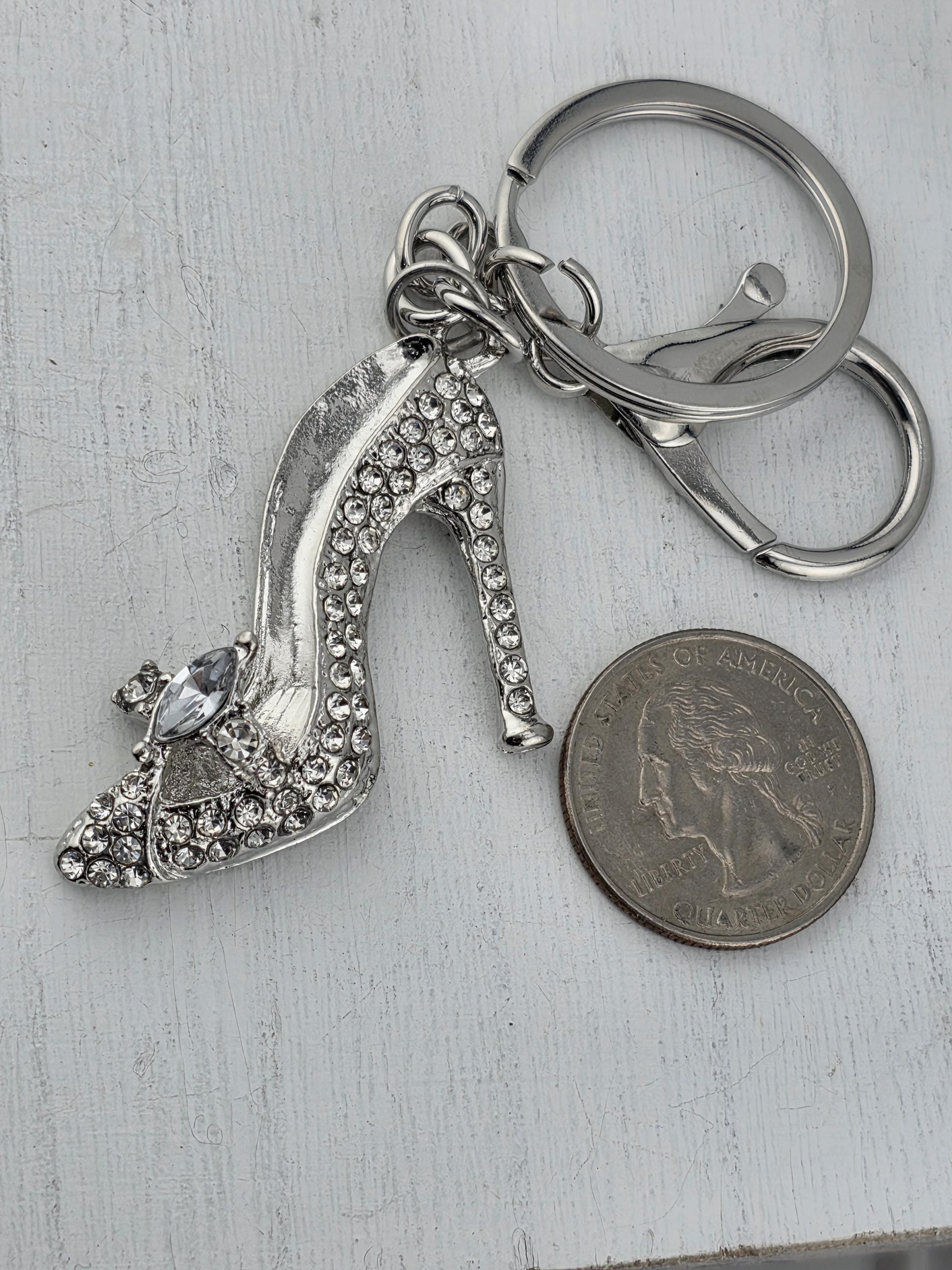 A silver keychain with a sparkling crystal heel shoe charm is placed next to a quarter coin on a white surface.