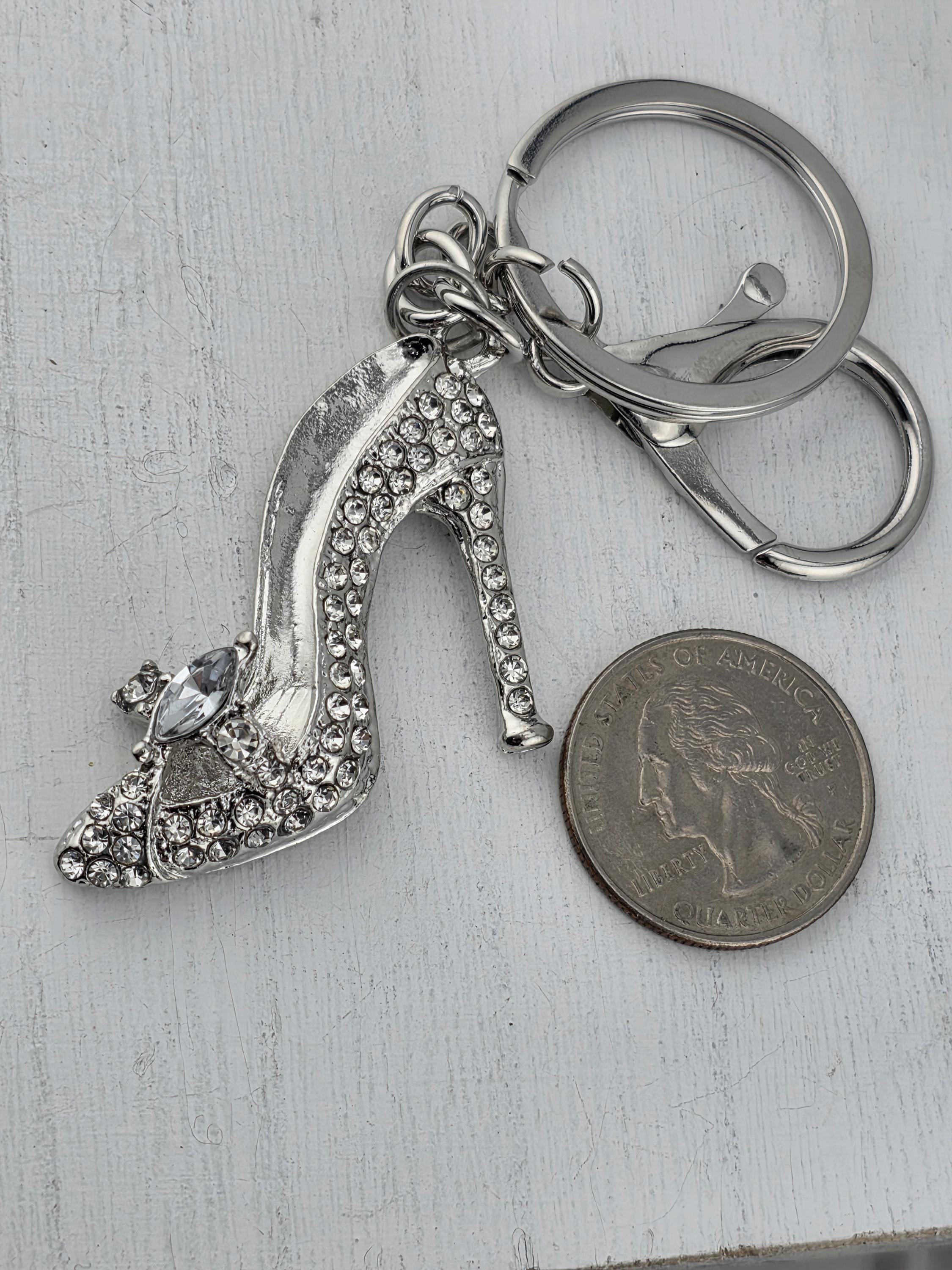 A silver keychain with a sparkling crystal heel shoe charm is placed next to a quarter coin on a white surface.