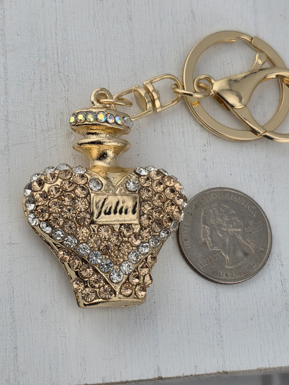 A decorative gold-colored perfume bottle with a heart-shaped design and sparkling rhinestones is displayed in a white ornate frame.