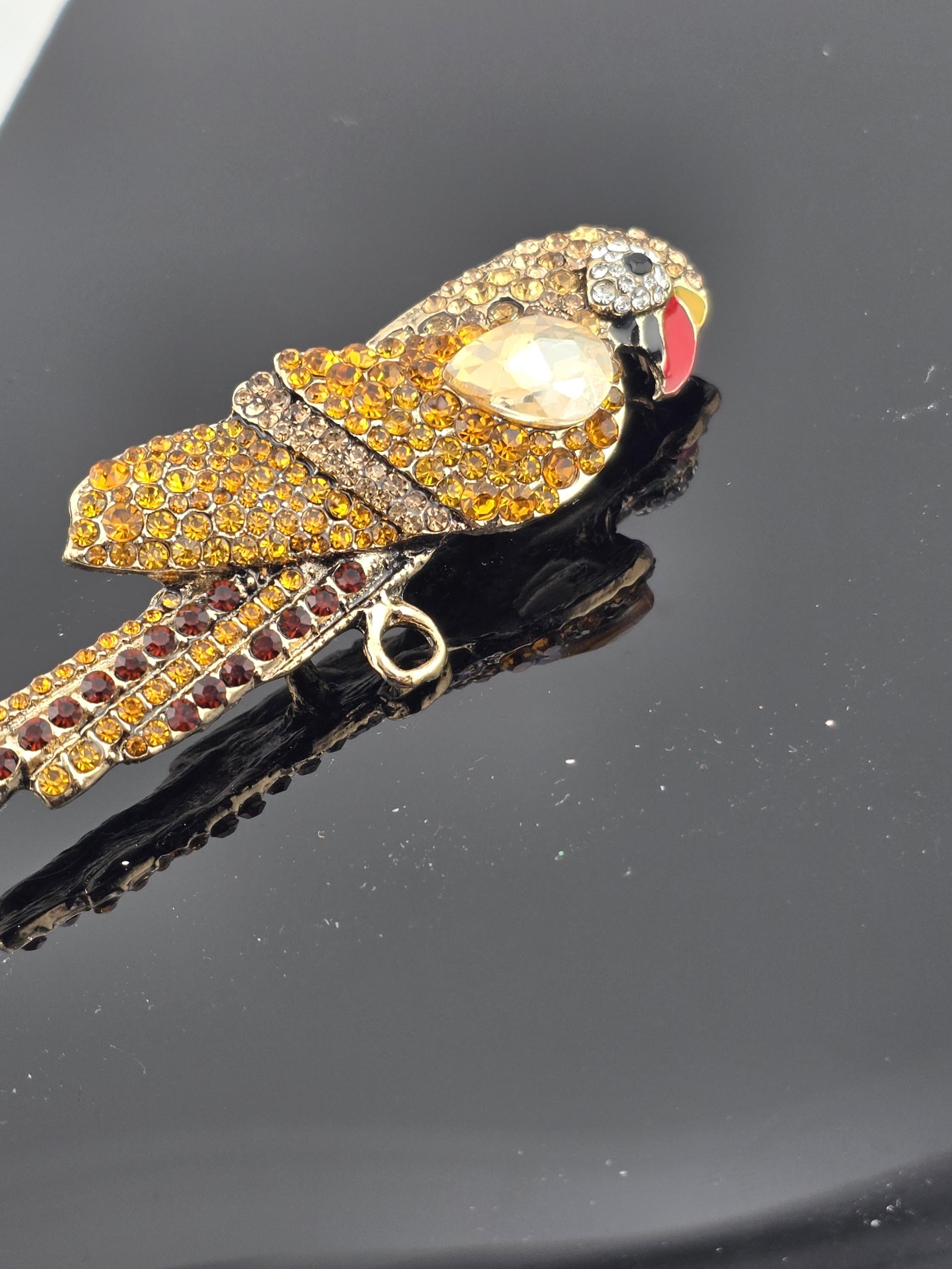 A decorative bird-shaped brooch with a yellow body, black wings, and a red beak, adorned with sparkling rhinestones and a large white gemstone.
