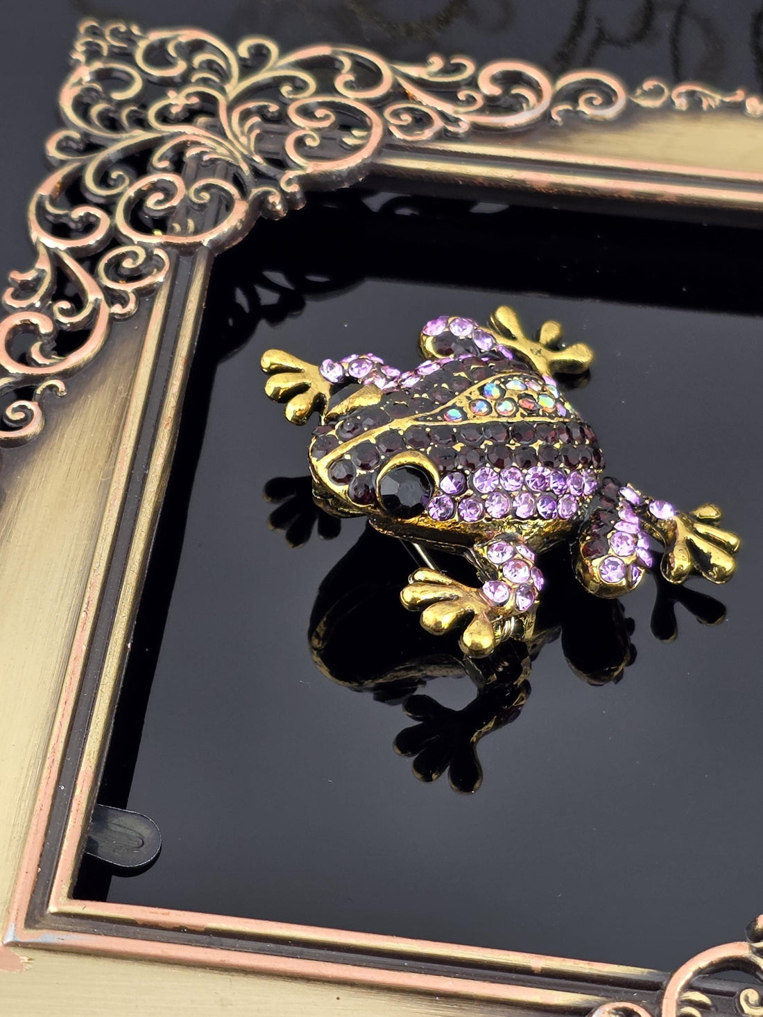 A decorative frog brooch with purple and gold accents, displayed in a gold frame with an ornate, scroll-like design.