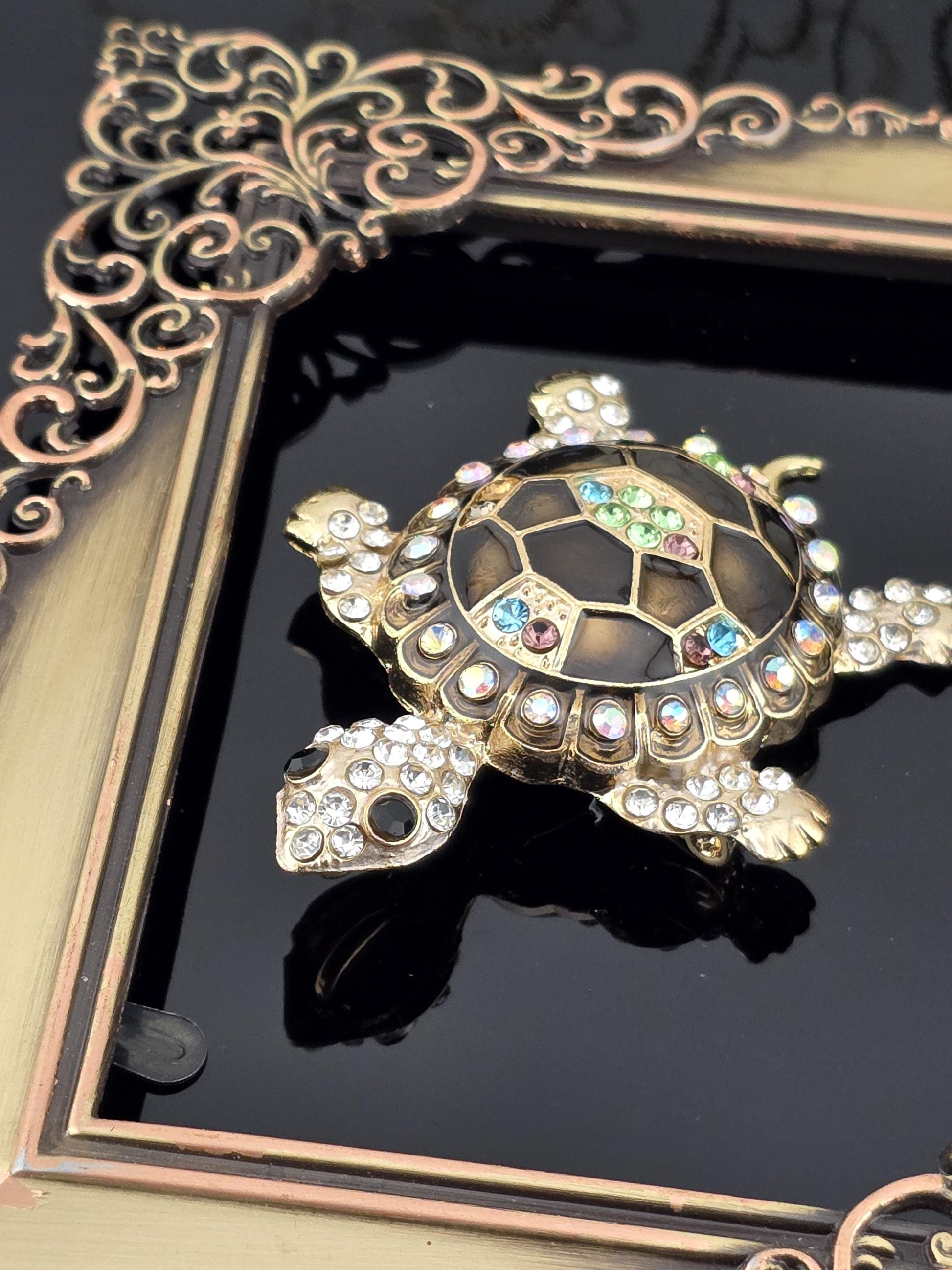 A decorative turtle-shaped brooch with a black background and a gold frame, adorned with colorful gemstones.