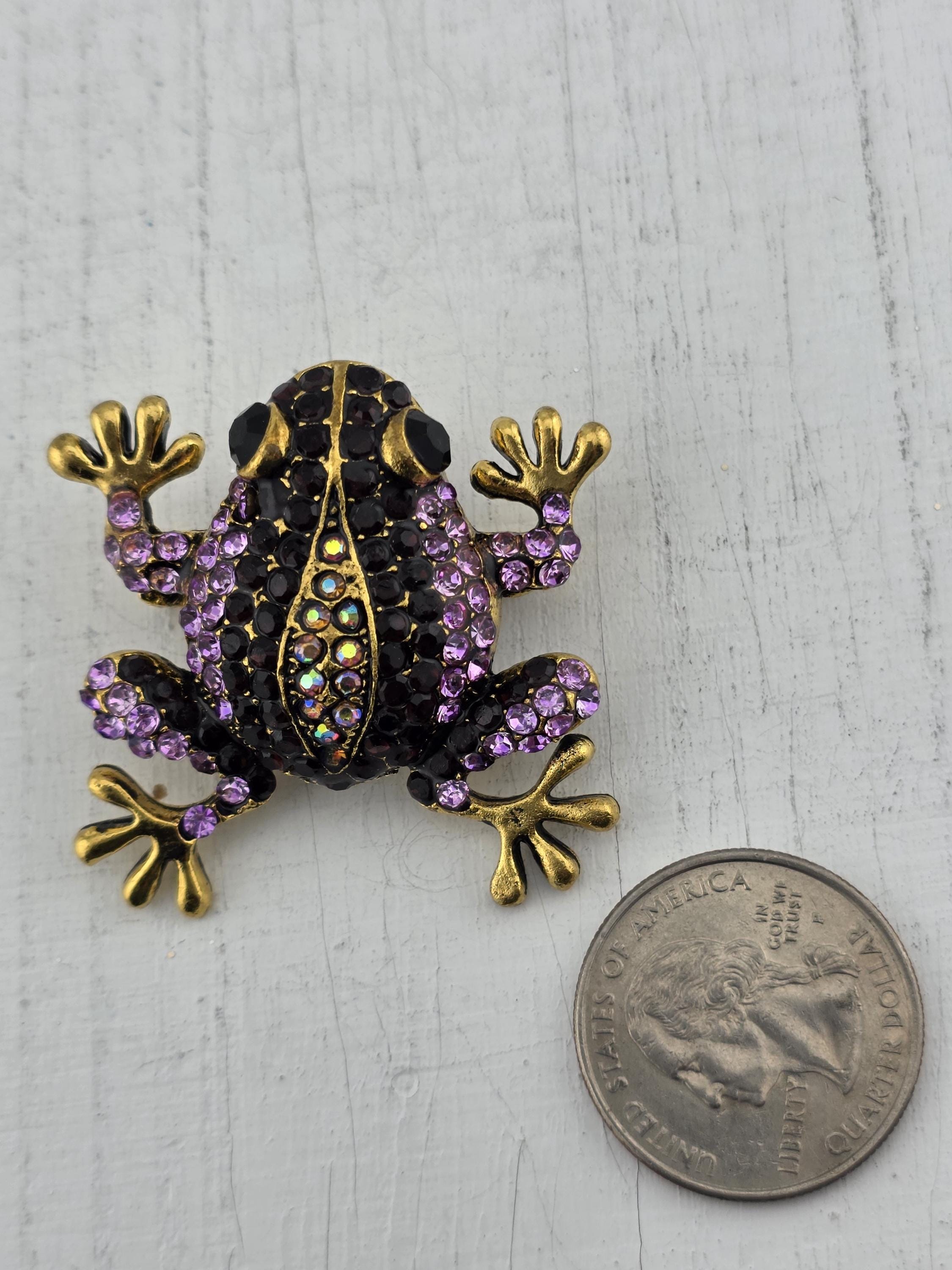 A decorative frog brooch with purple and gold accents, adorned with sparkling jewels, is placed next to a United States quarter on a white surface.