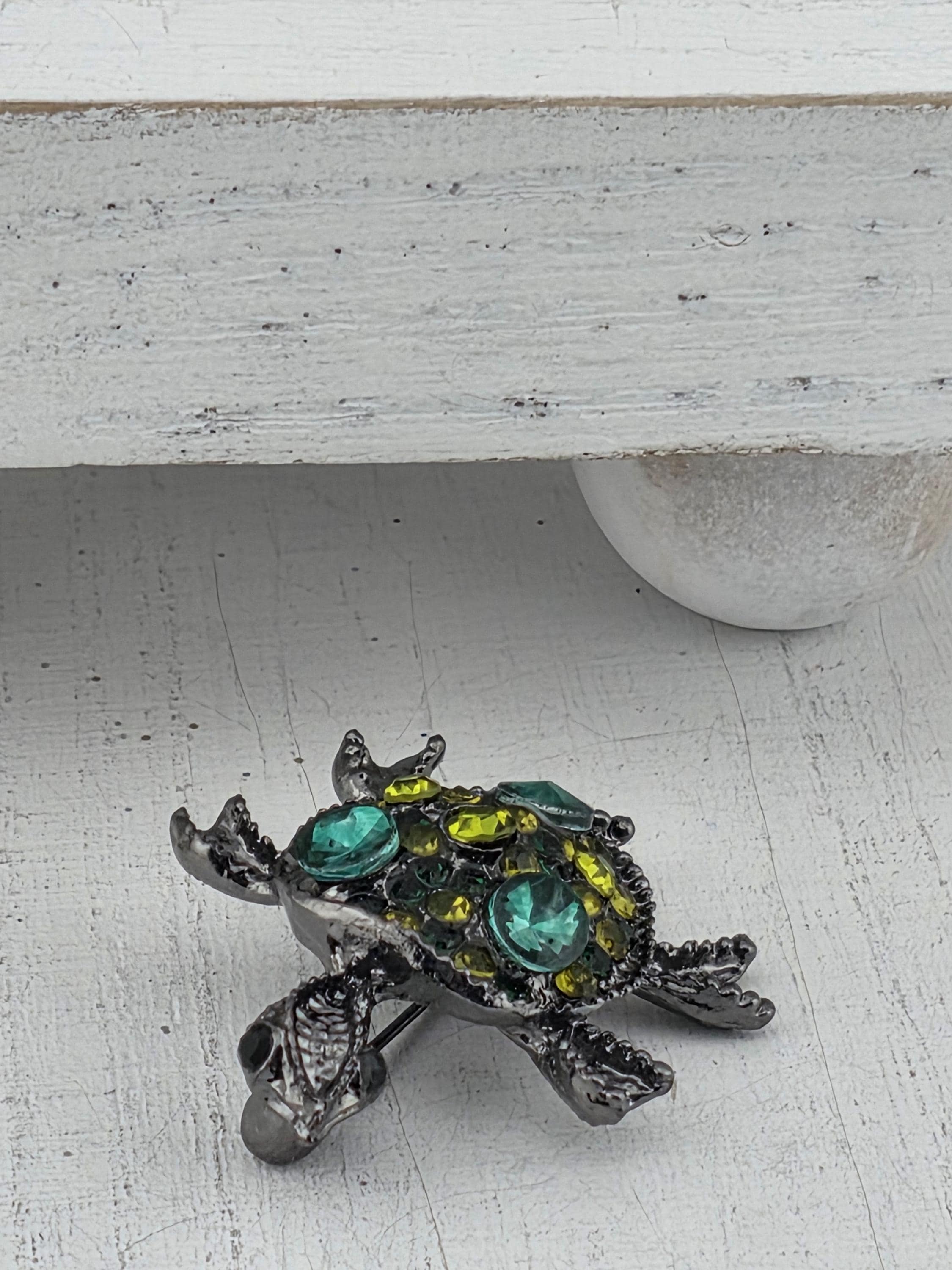 A decorative metal turtle brooch with green and yellow gemstones is displayed on a white wall.