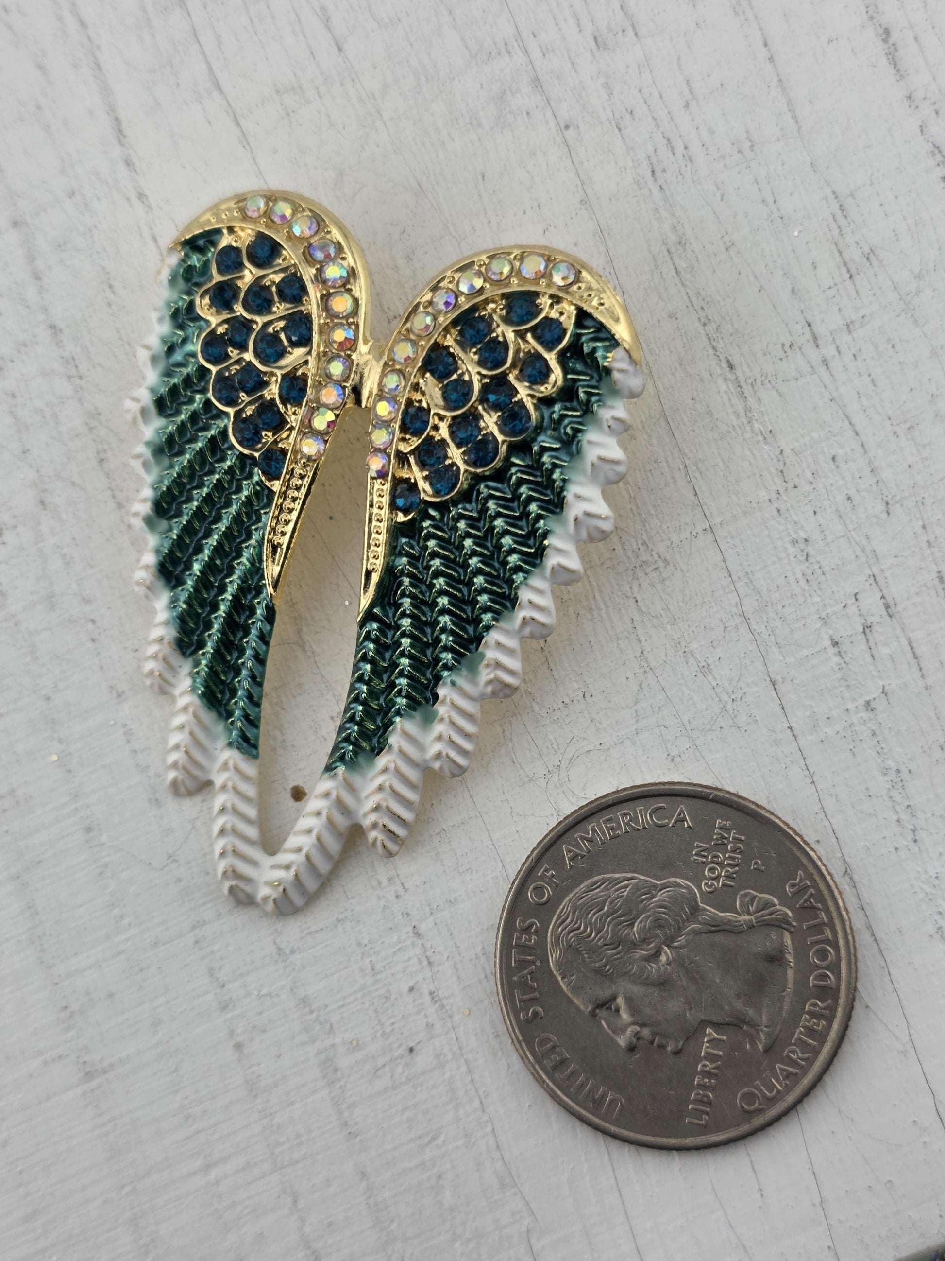 a decorative wing-shaped brooch with green and blue gemstones, placed next to a United States quarter coin.