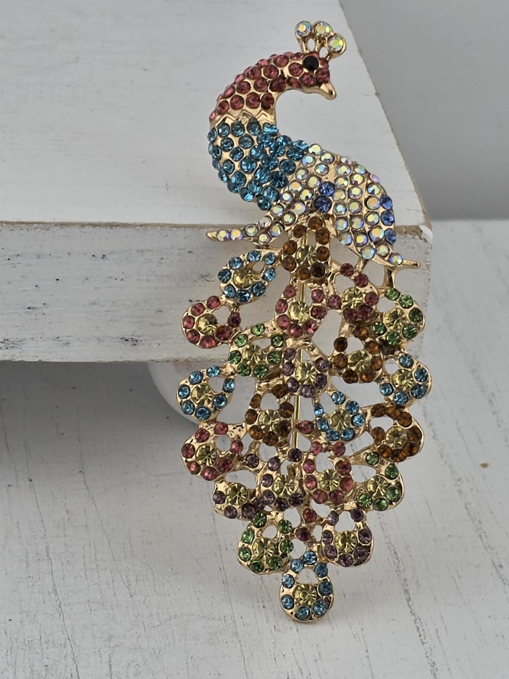 A decorative peacock brooch with a vibrant array of colorful gemstones, including blue, green, red, and yellow stones, set against a white background.