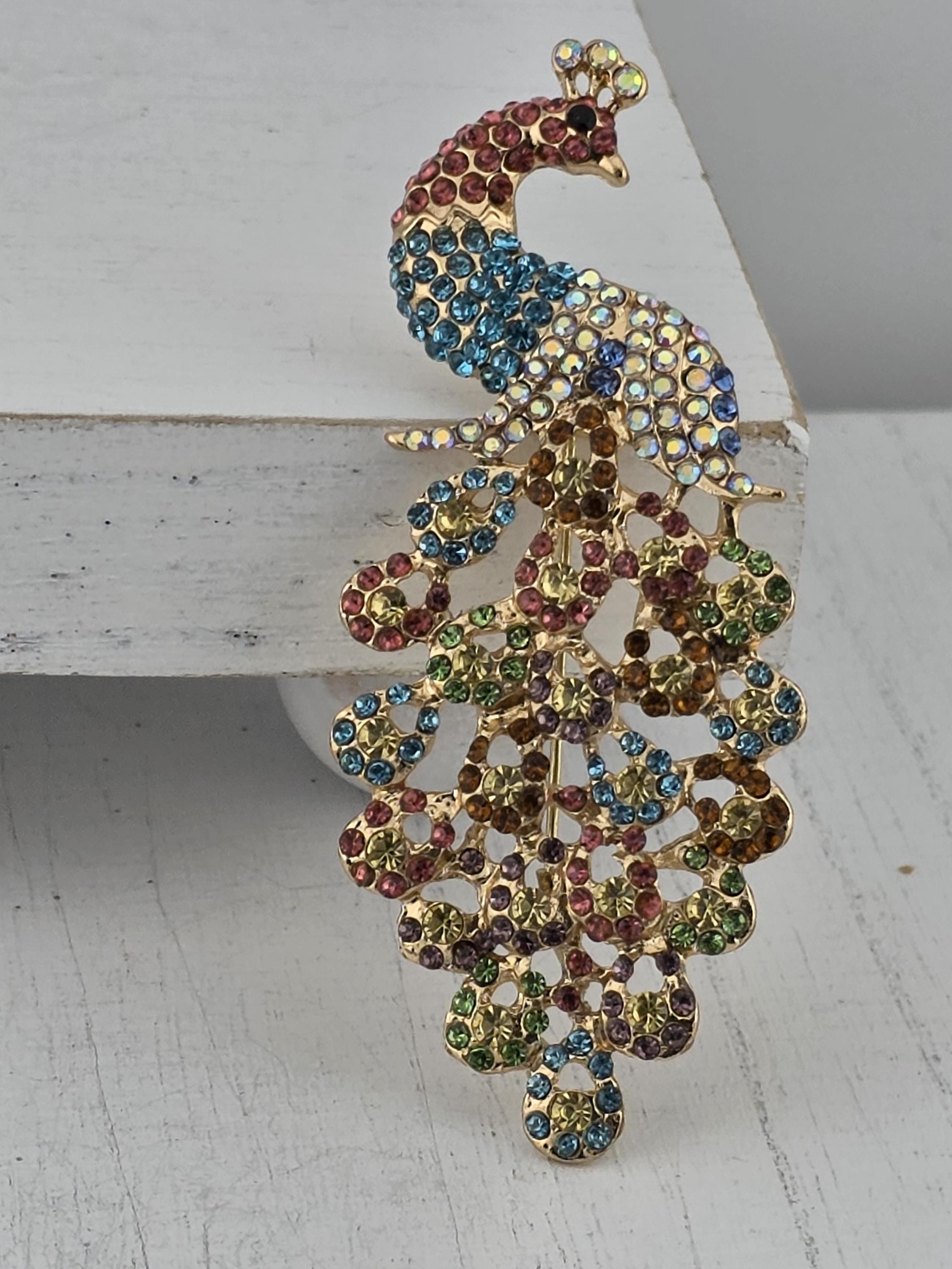 A decorative peacock brooch with a vibrant array of colorful gemstones, including blue, green, red, and yellow stones, set against a white background.