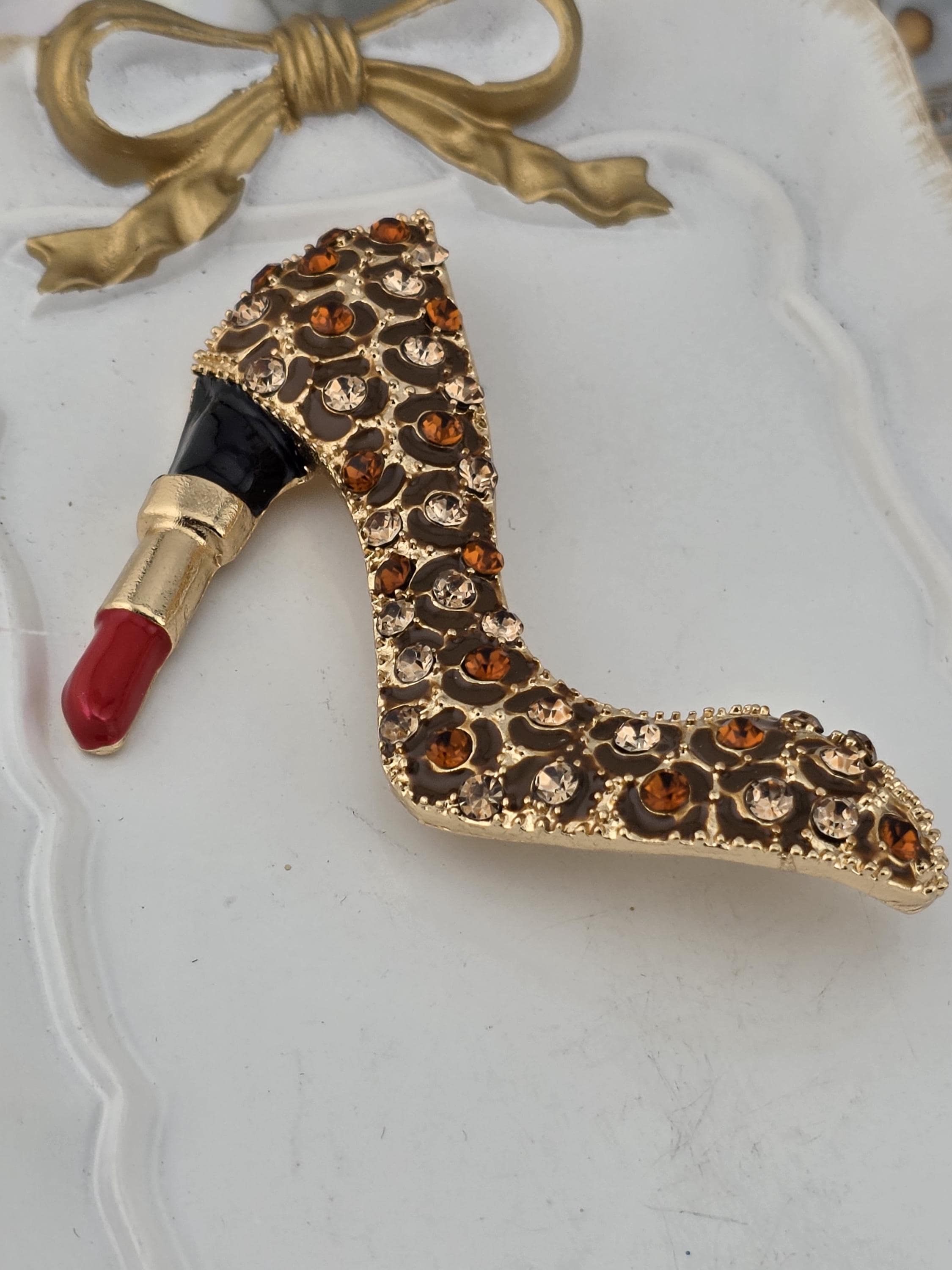 A gold and red high heel shoe brooch with a red lipstick cap is displayed on a white surface.