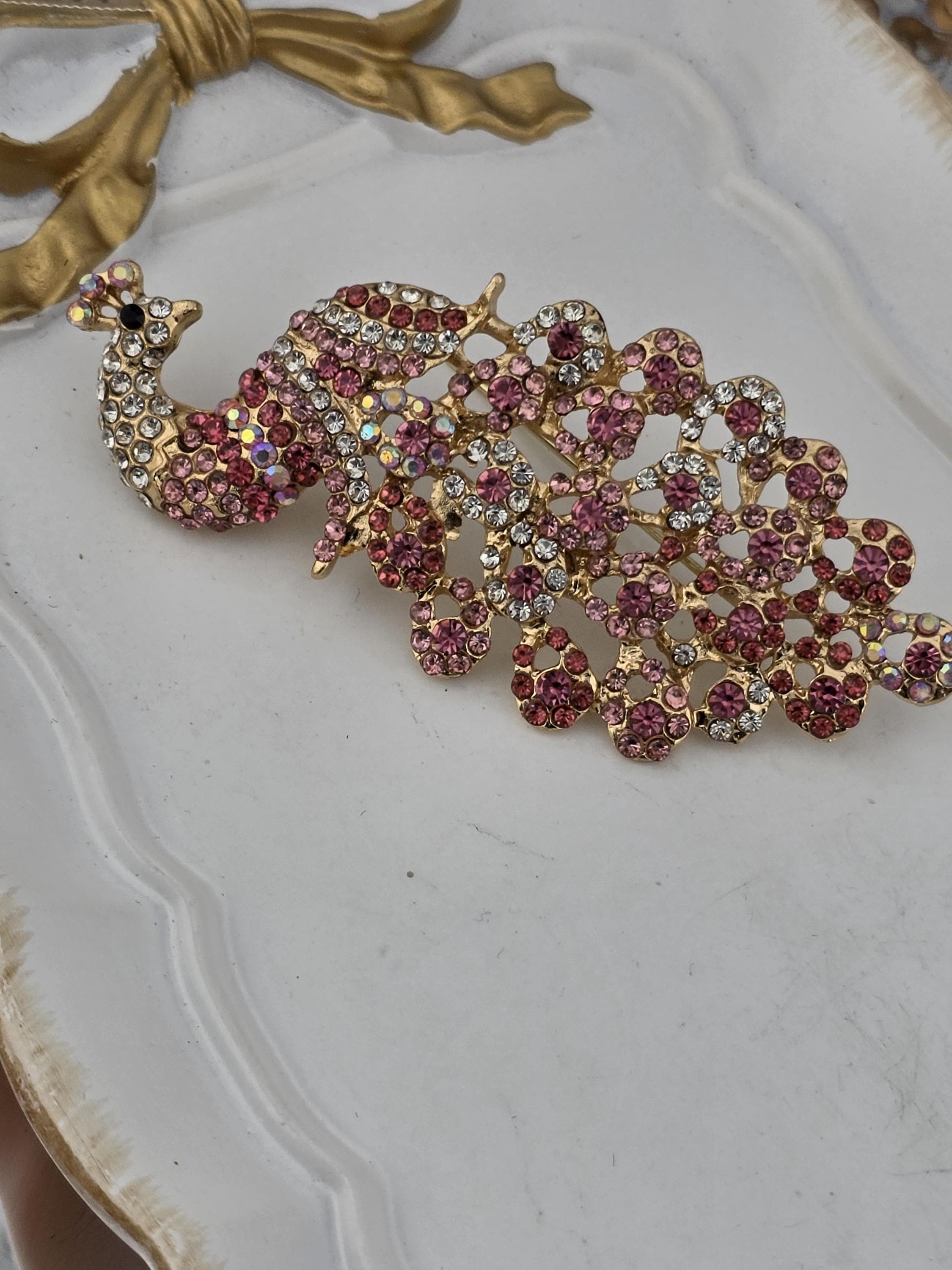 A decorative peacock brooch with a pink and gold design, featuring numerous small gemstones.