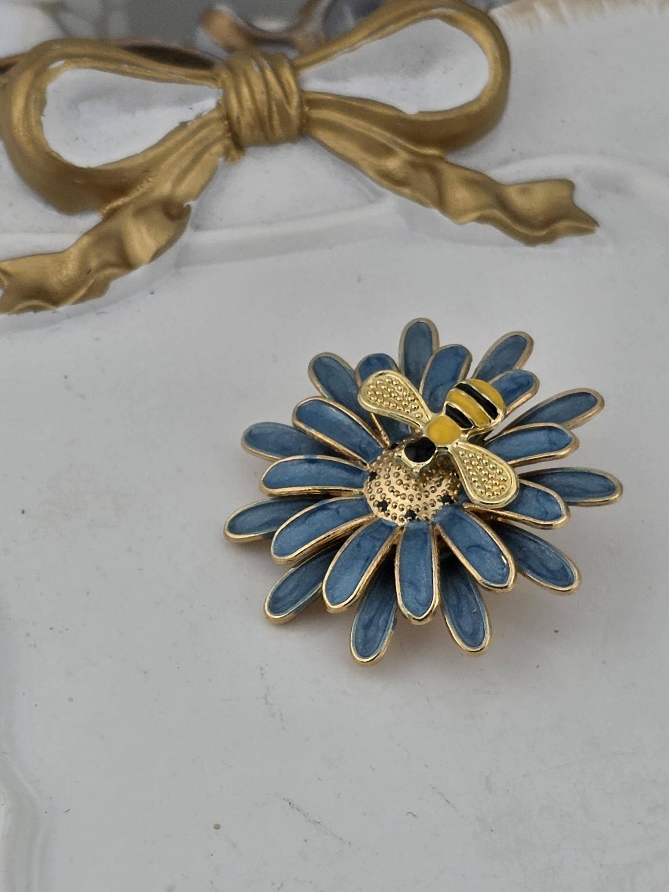 A decorative blue flower brooch with a gold bee on it, placed on a white surface.