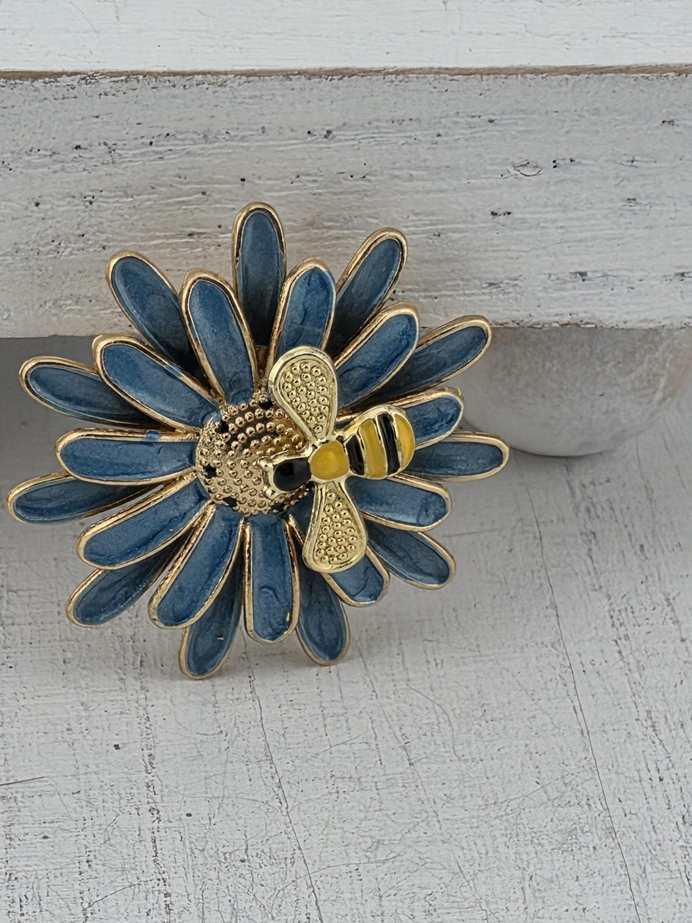 A decorative blue and gold flower-shaped brooch with a bee on it, placed on a white wall.