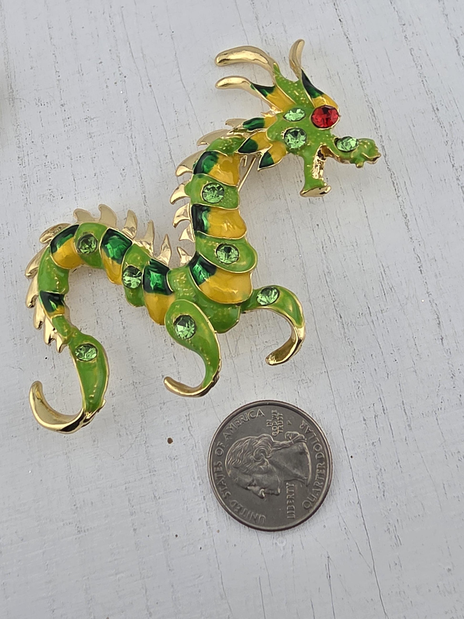 A colorful dragon brooch with green and yellow scales and a red gem on its head is placed next to a coin on a white surface.