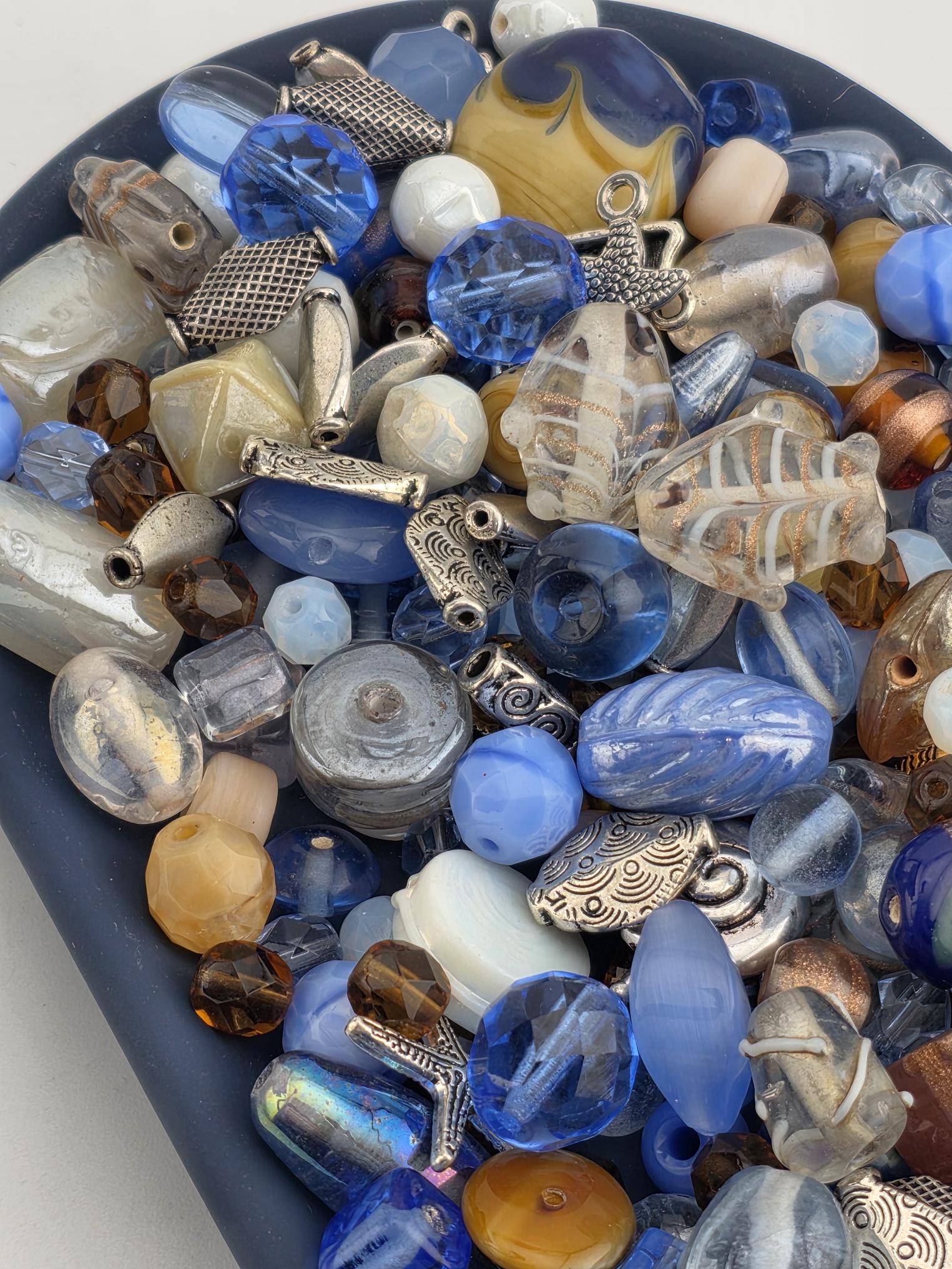 A collection of various beads, charms, and glass objects in shades of blue, silver, and brown, arranged on a surface.