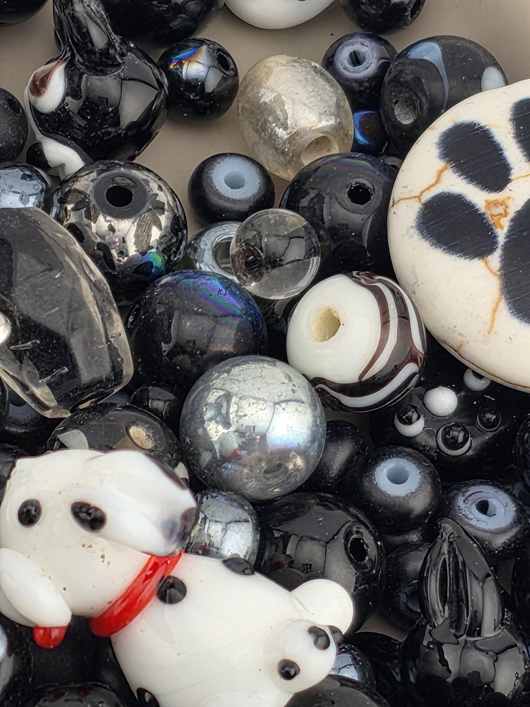 The image is a close-up of a collection of various beads and glass objects, including a large white and black spotted object, a small white and black striped object, and several black and silver objects.