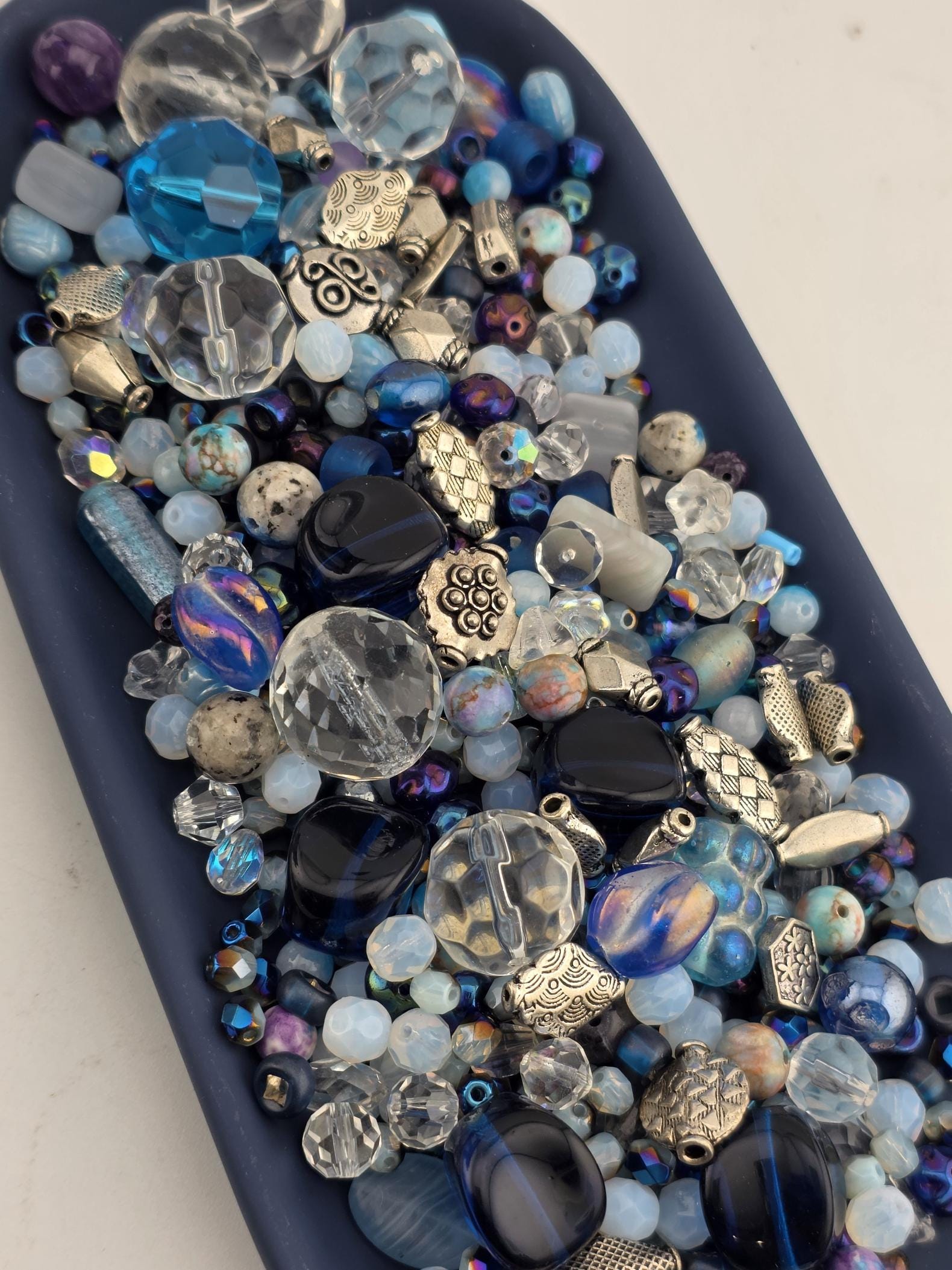 A collection of various colored glass beads and metal pieces, including clear, blue, and silver beads, arranged on a dark surface.