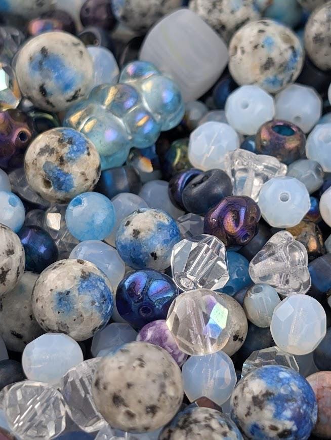 The image is a close-up of various beads and gemstones, including clear, blue, purple, and white beads, as well as some larger stones.