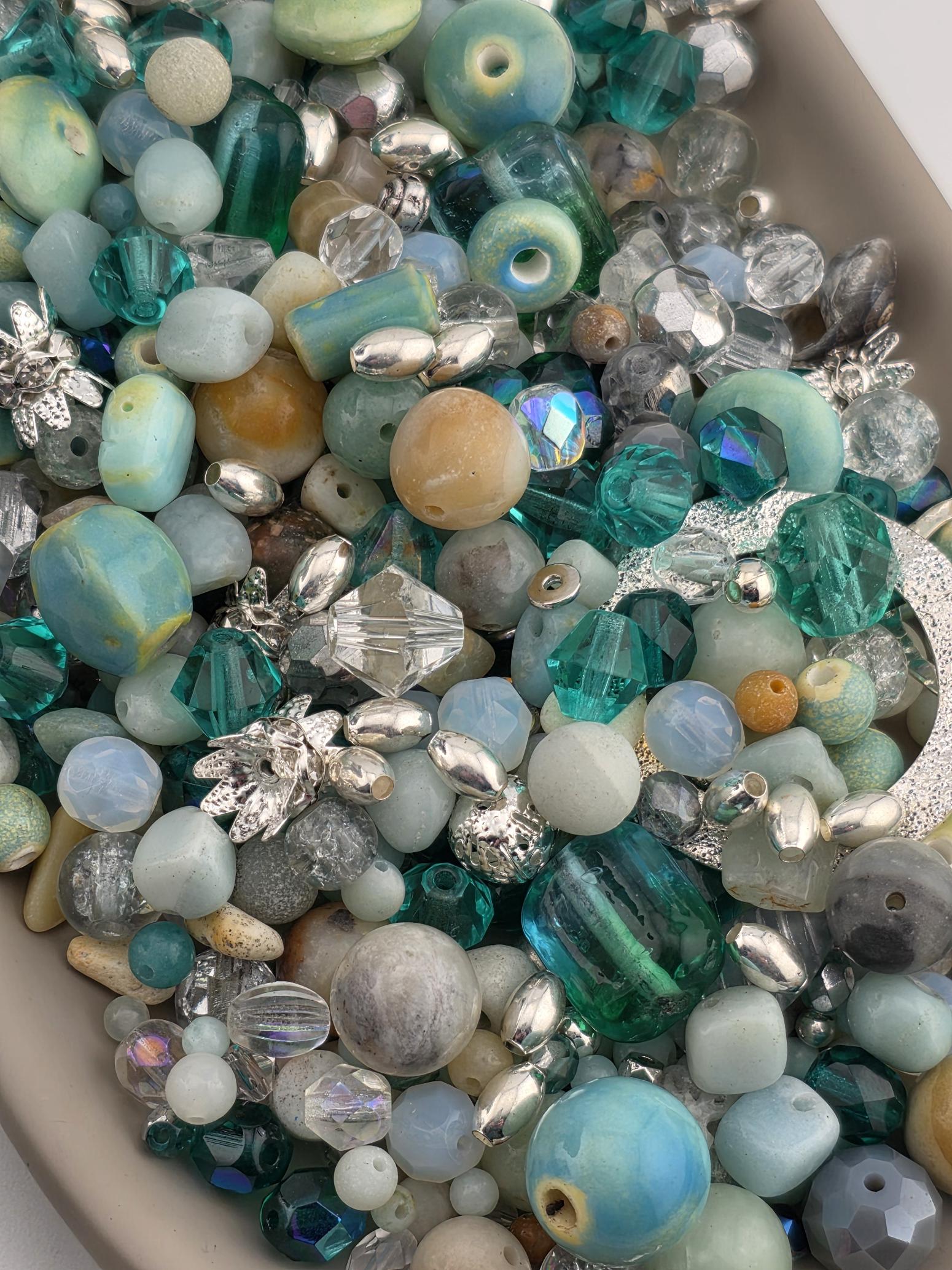 The image is a close-up of a collection of various beads and gemstones, including clear, blue, green, and silver beads, as well as some larger stones.