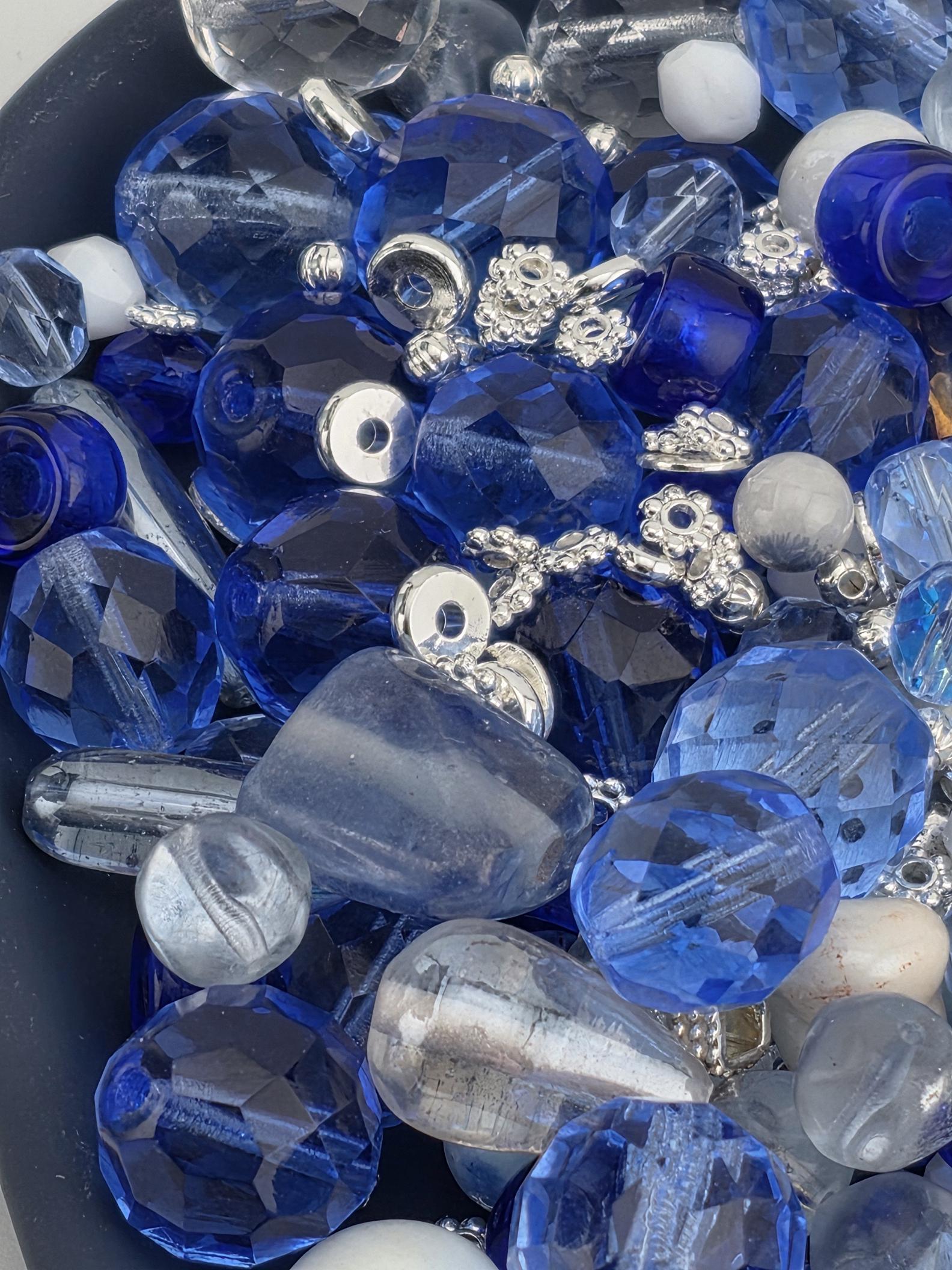 A close-up image of various blue and silver glass beads and charms, including clear, blue, and silver beads, as well as some silver and blue glass beads.