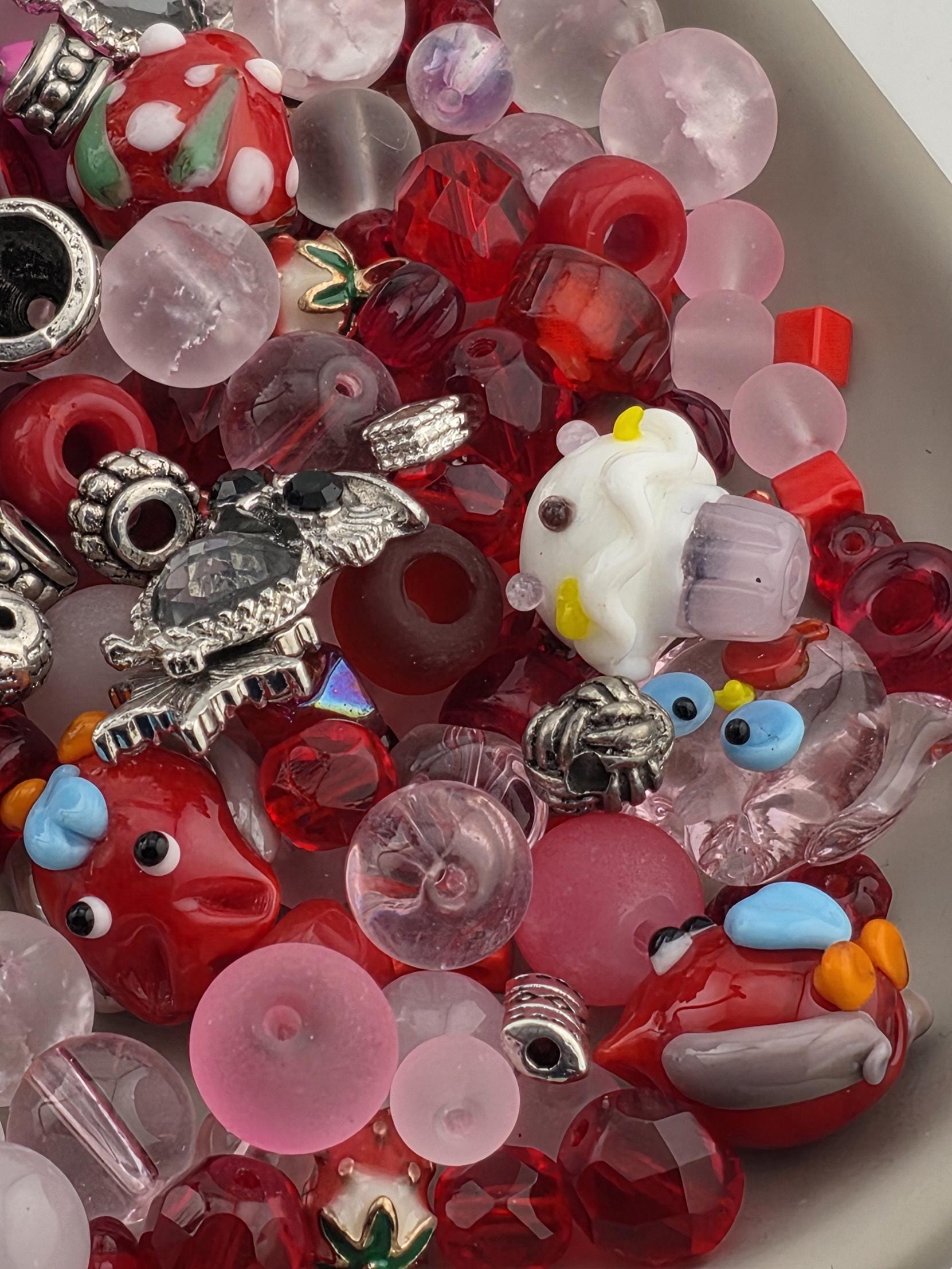 The image is a colorful assortment of various beads, charms, and glass objects, including clear and red glass beads, metal charms, and other decorative items.