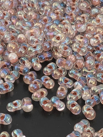 a close-up view of numerous small, clear beads scattered across a dark surface. The beads appear to be made of glass and have a transparent, iridescent quality, reflecting light in various colors. The image does not contain any text or specific details about the beads, and it does not depict any actions or movements. The focus is on the individual beads and their arrangement on the surface.