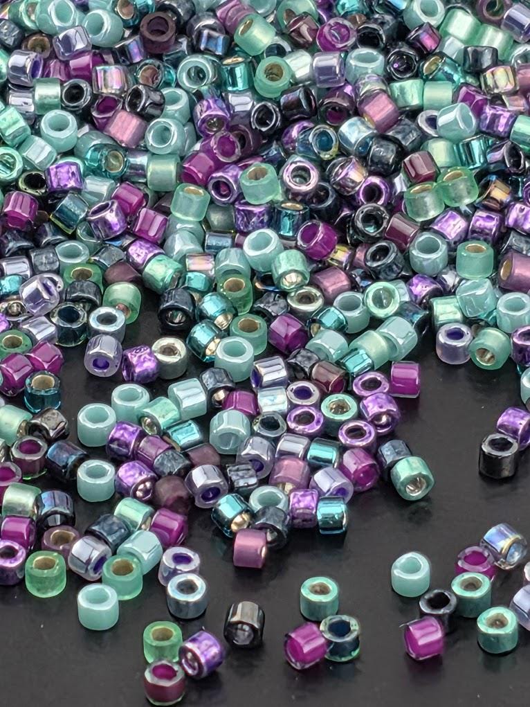 A colorful assortment of multicolored beads, including shades of purple, green, blue, and silver, scattered across a dark background.