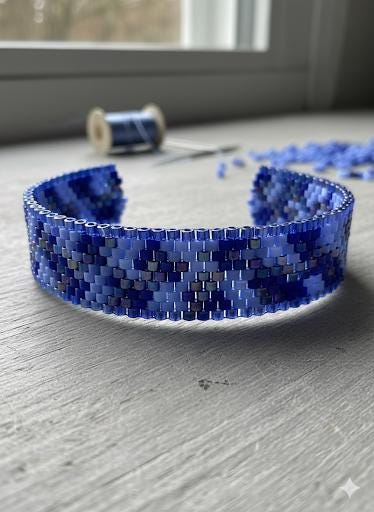 A blue and white beaded bracelet is displayed on a wooden surface, with a spool of thread and scattered beads visible in the background.