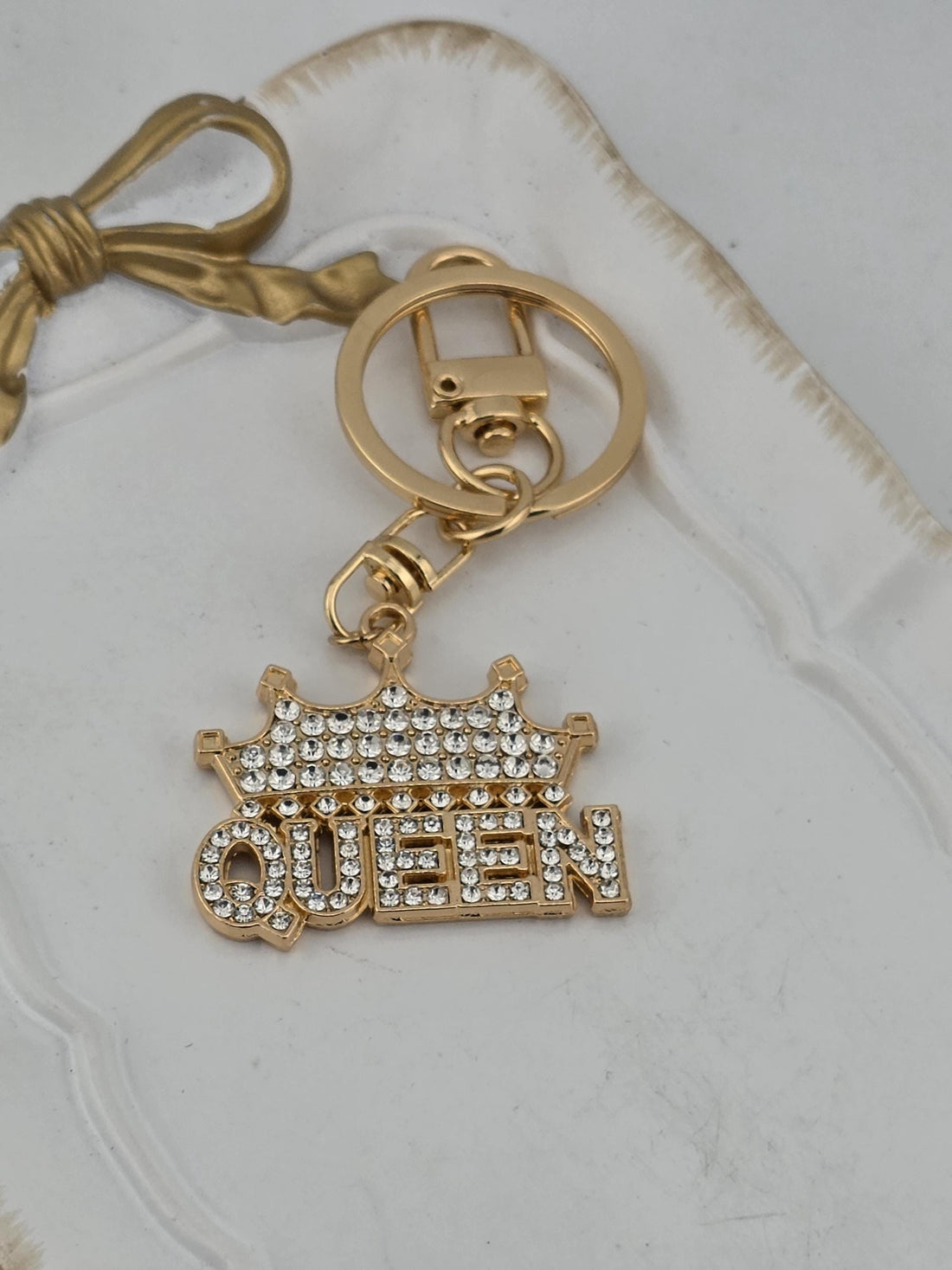 A gold-colored keychain with a charm that says "QUEEN" in a decorative font, made of small rhinestones or crystals.
